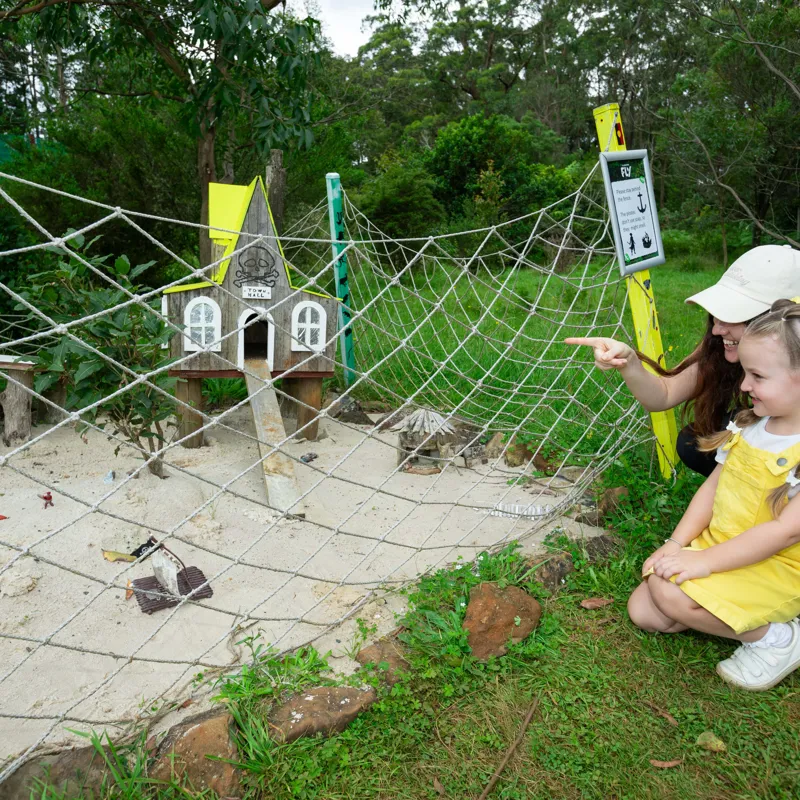 Two children, a woman and a girl, look at a small birdhouse designed to look like a haunted house with a spooky skull sign on top, surrounded by a netted barrier, in a park or natural area.