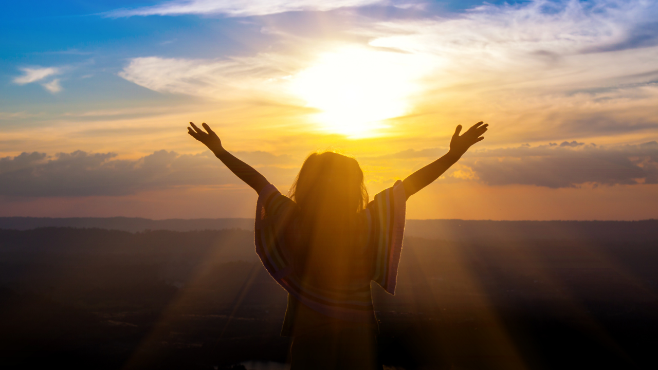 Person standing outdoors with arms raised during sunset, silhouette against a colorful sky with clouds.