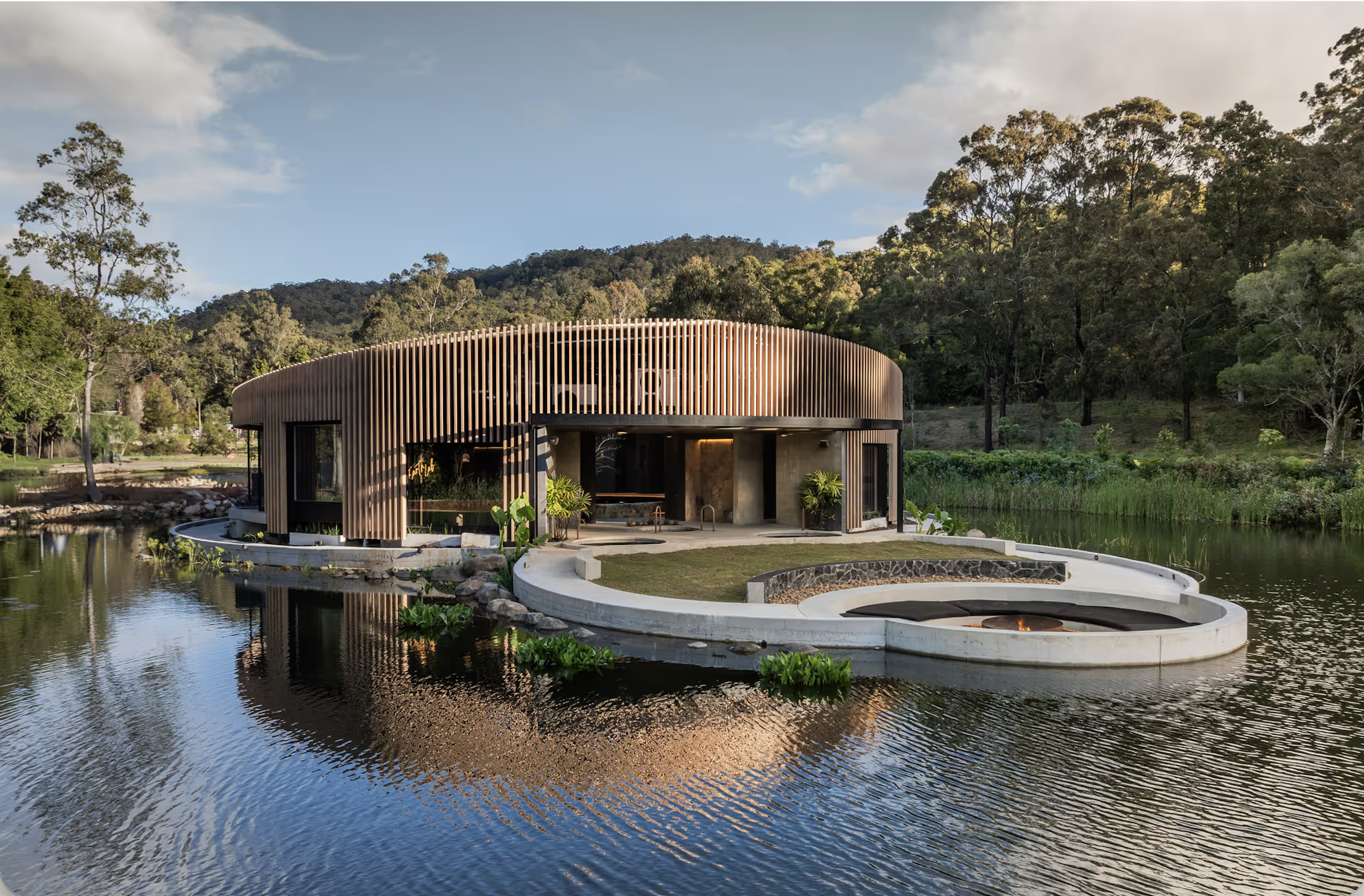 Modern house with wooden vertical slats, surrounded by water and green trees, with a fire pit and landscaped yard.