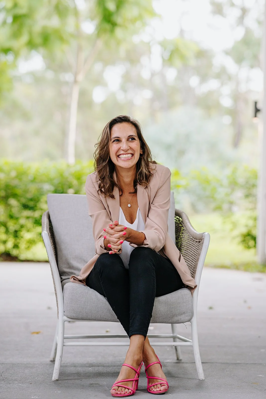 A woman sitting on a light gray outdoor chair, smiling with her hands clasped, wearing a beige blazer, white top, black pants, and pink high-heeled sandals, outdoors with green trees in the background.