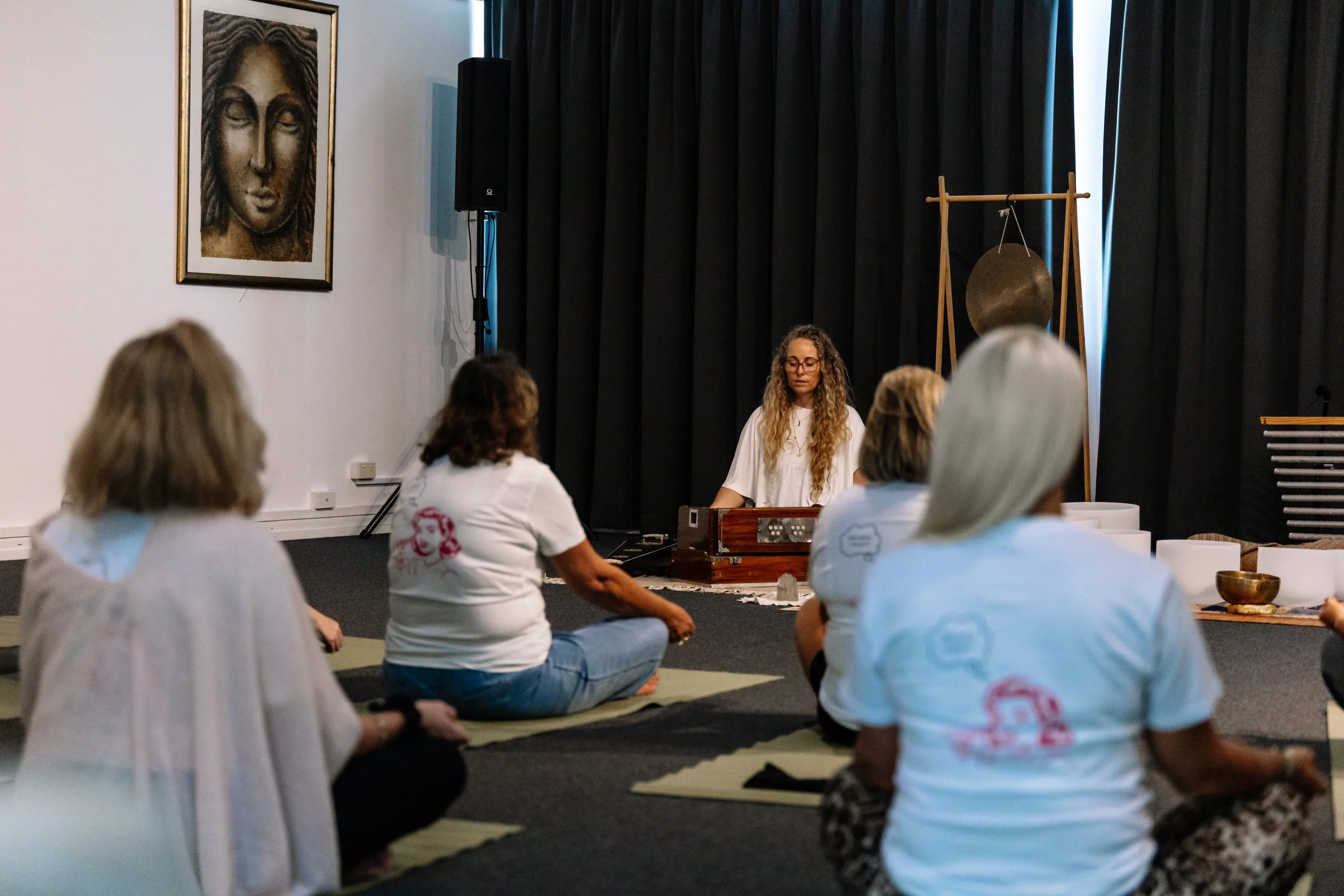 A woman with long curly hair and glasses sits on the floor in front of a small group, playing a wooden instrument. The group is sitting on mats, facing her, in a room with black curtains and various meditation or sound therapy items, including singin