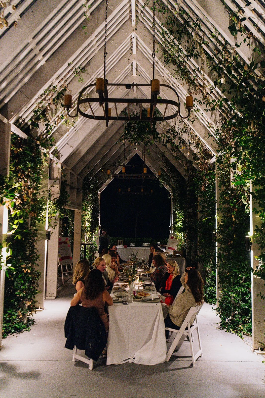 A group of people dining at a long table under a decorative, lit garden-themed pavilion at night.