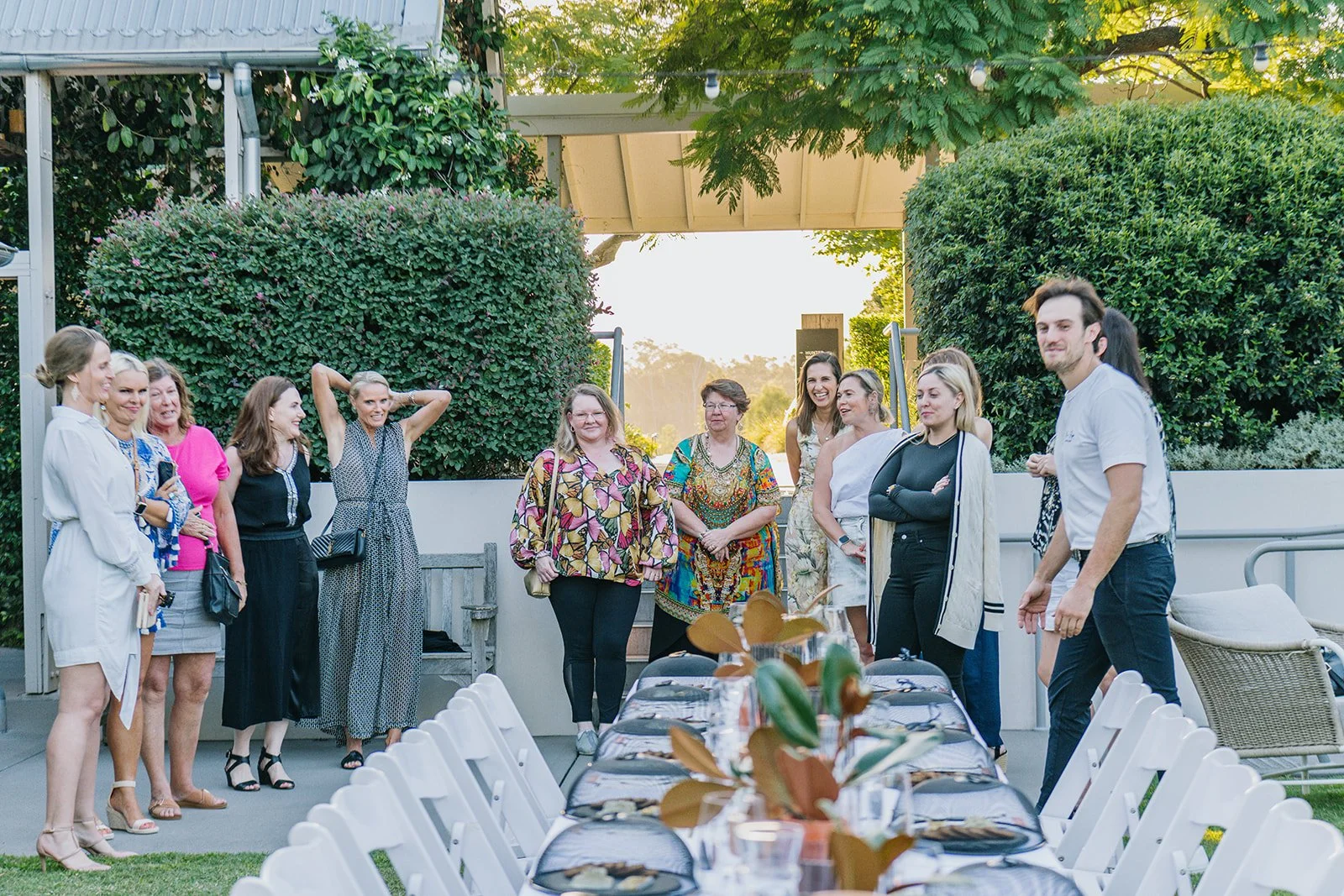 A group of people gathered outdoors around a long table with plates, glasses, and decorations, smiling and engaging in conversation during a social event.