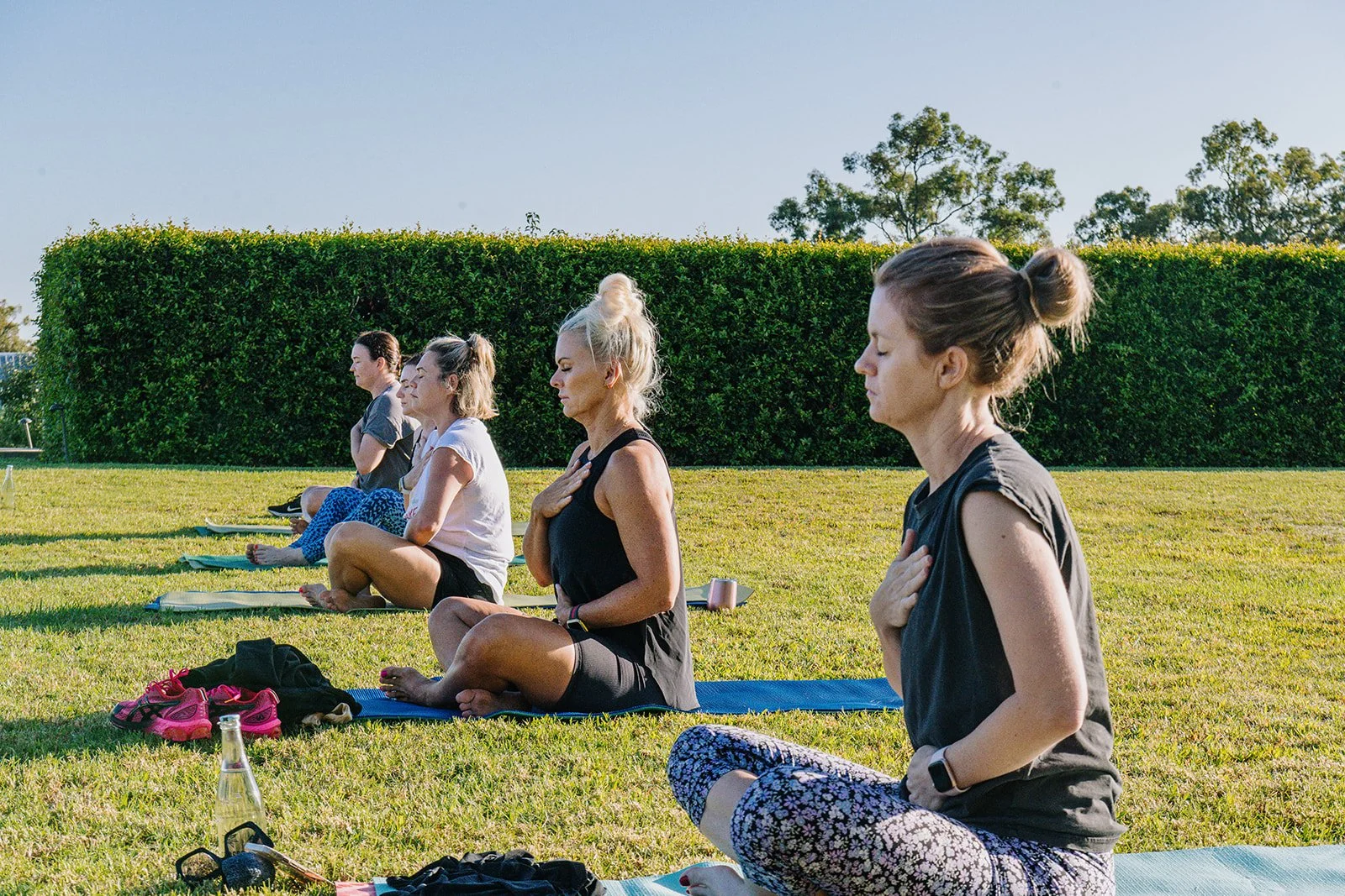 Women practicing yoga outdoors on mats in a grassy field with a background of a large green hedge and clear sky.