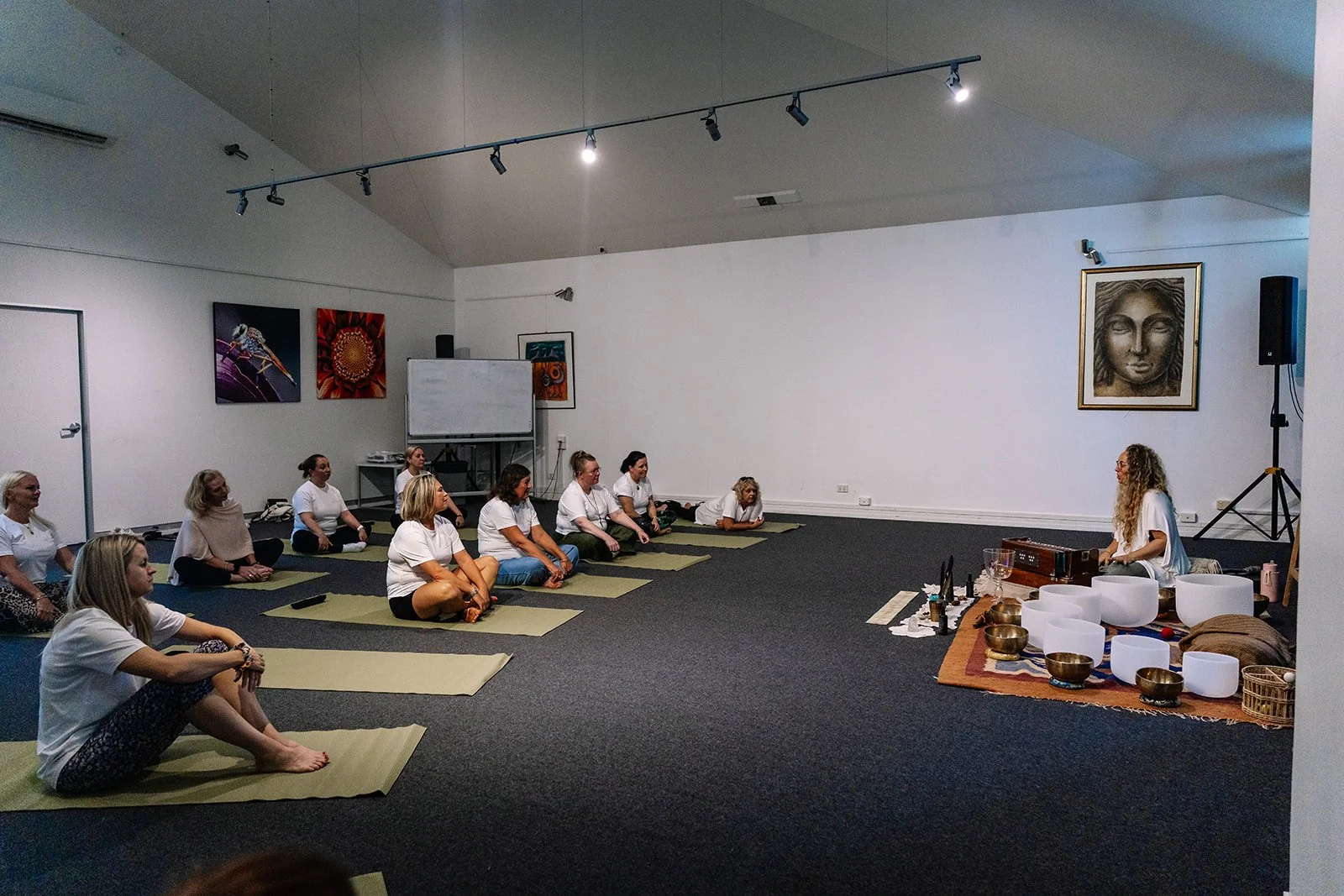 A group of women participating in a yoga or meditation class in a room, sitting on mats, facing a woman sitting on a mat with singing bowls and other sound therapy instruments, with artwork on the walls.