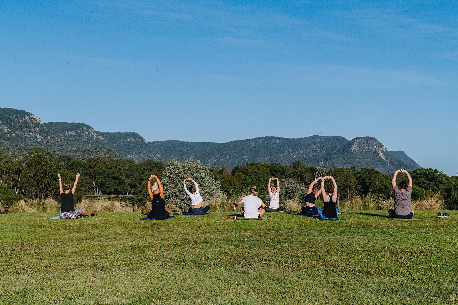 Group of people practicing yoga outdoors on a grassy field with mountains and blue sky in the background.