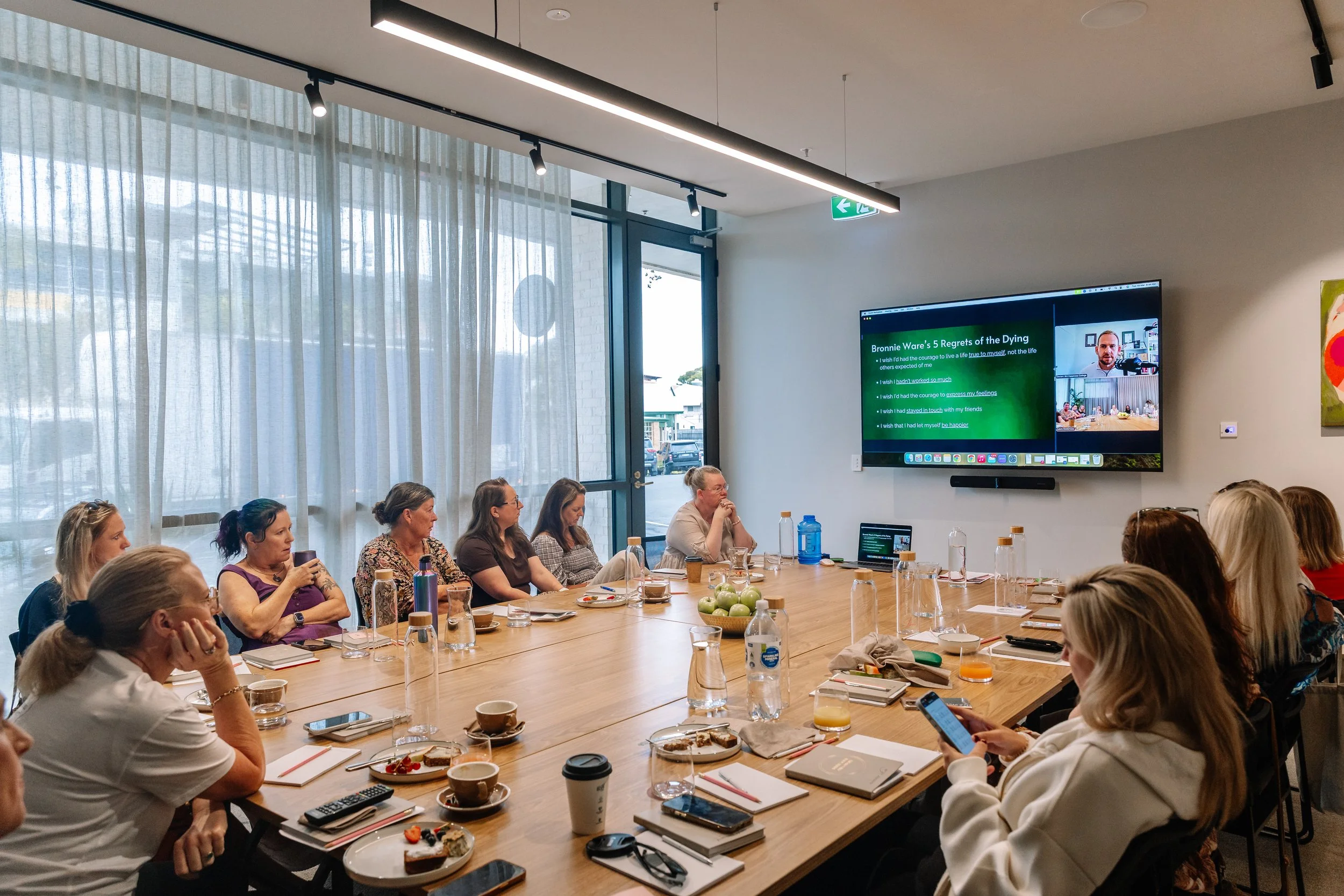 A group of women attending a meeting in a modern conference room with a large window, a big screen displaying a presentation, and various notebooks and beverages on the table.