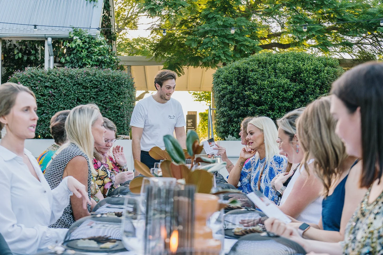 A group of women sit at a long outdoor dining table, engaged in a conversation, with a man standing and speaking, surrounded by greenery and string lights.