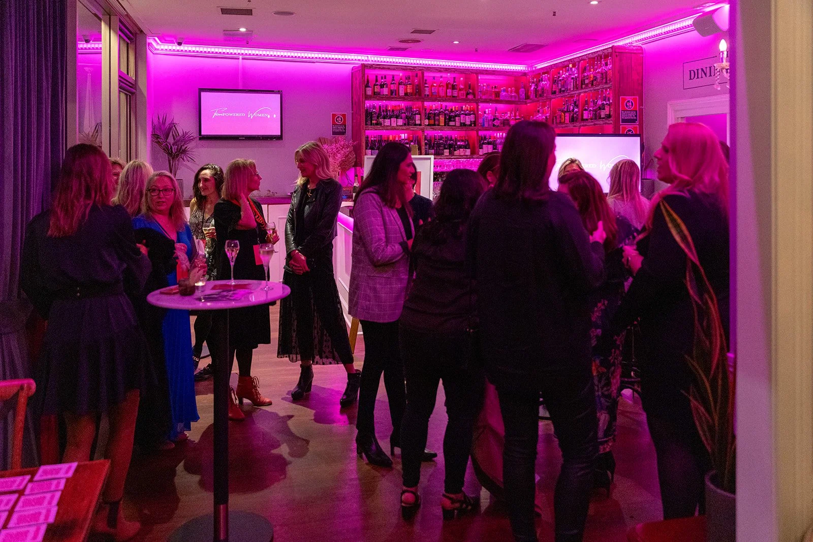 Group of women at a social gathering in a bar with pink neon lighting, bottles on shelves, and a TV screen displaying 'Empowered Women'.