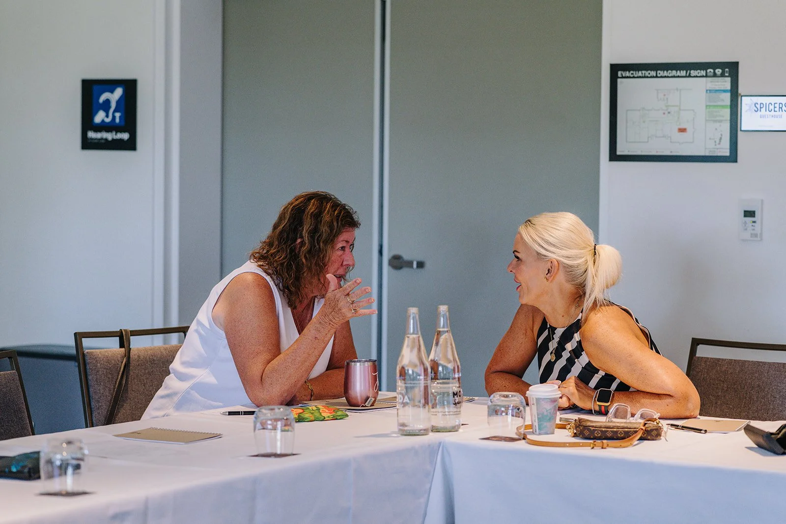 Two women sitting at a conference table engaged in a lively conversation, smiling and gesturing, with water bottles, a coffee cup, and notebooks on the table, in a meeting room with informational signs on the wall.
