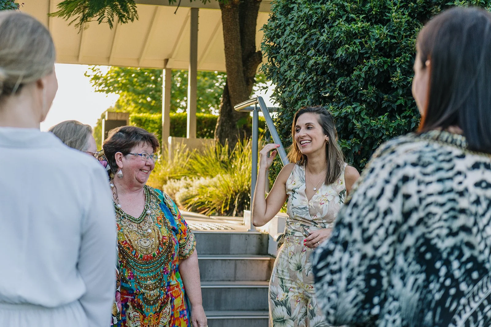 Group of women smiling and talking outdoors near a staircase with greenery and trees in the background.