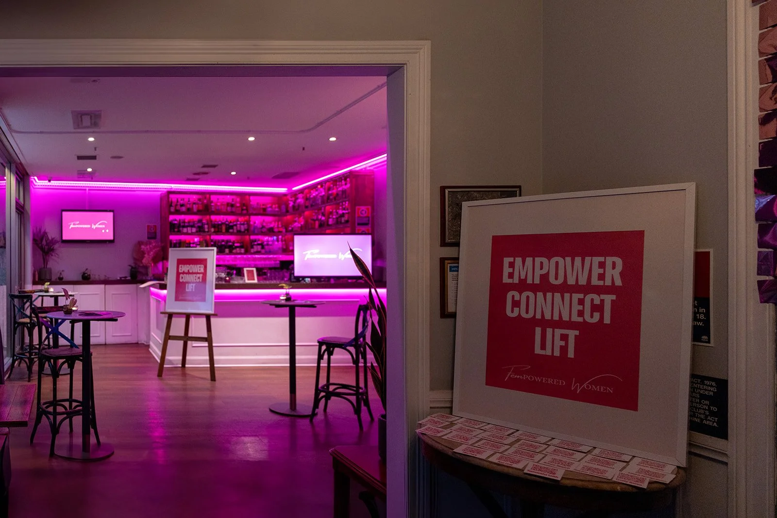 Interior of a room with pink neon lighting, a bar with bottles on shelves, and a sign that reads 'Empower Connect Lift' in front of the bar.