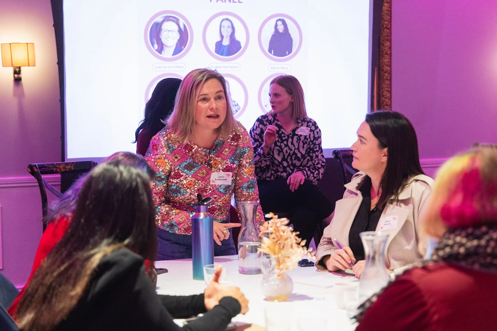 Women participating in a panel discussion at a conference, seated around a table with water bottles and a vase with flowers, with a presentation slide in the background showing headshots of women.