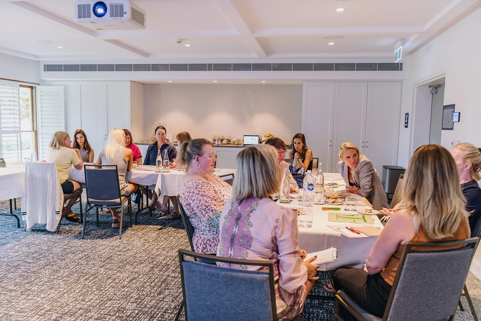 A group of women seated around a conference table engaged in a meeting in a well-lit room with white walls, cabinets, and windows with shutters.