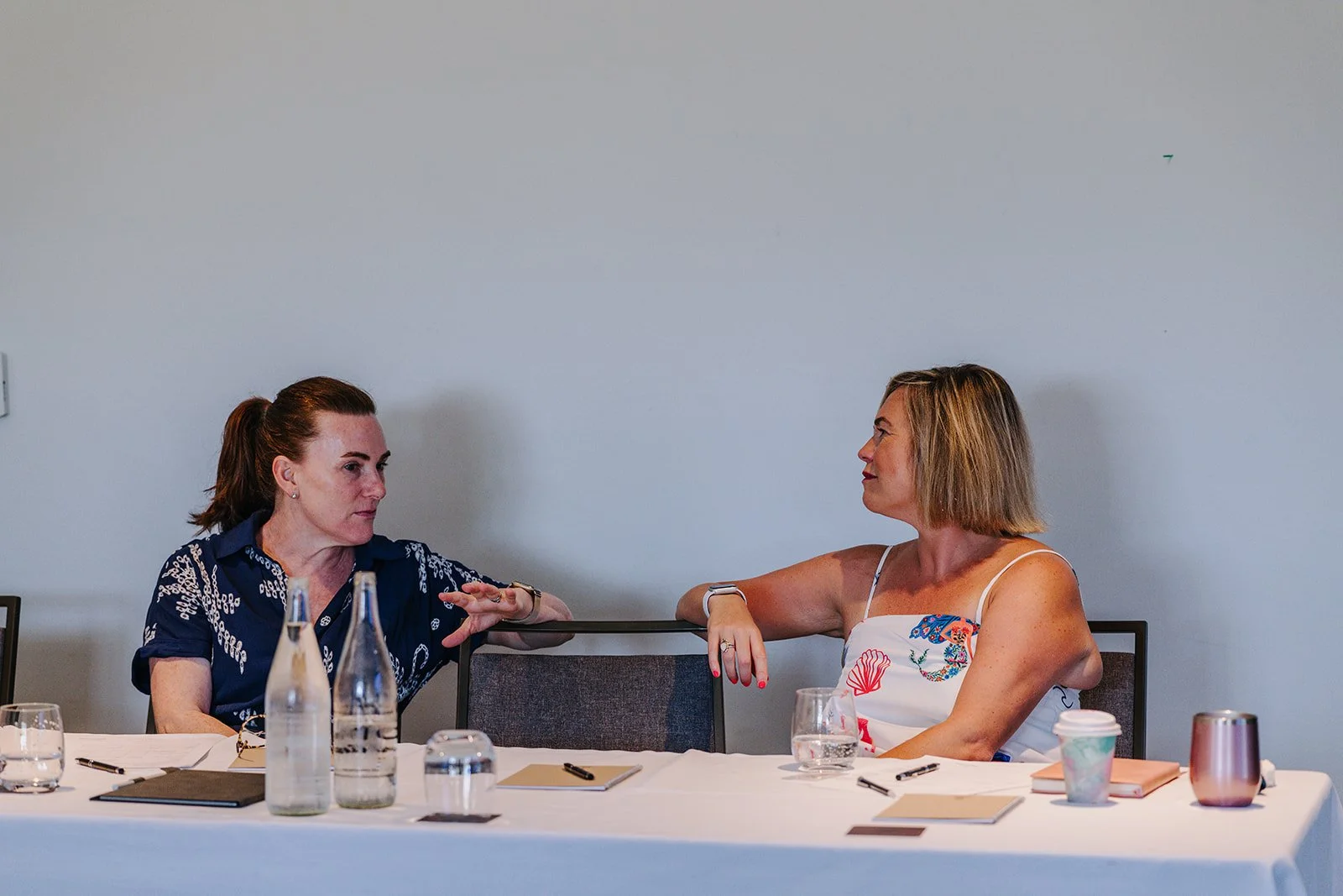 Two women sitting at a conference table having a discussion, with water bottles, notebooks, and coffee cups on the table.