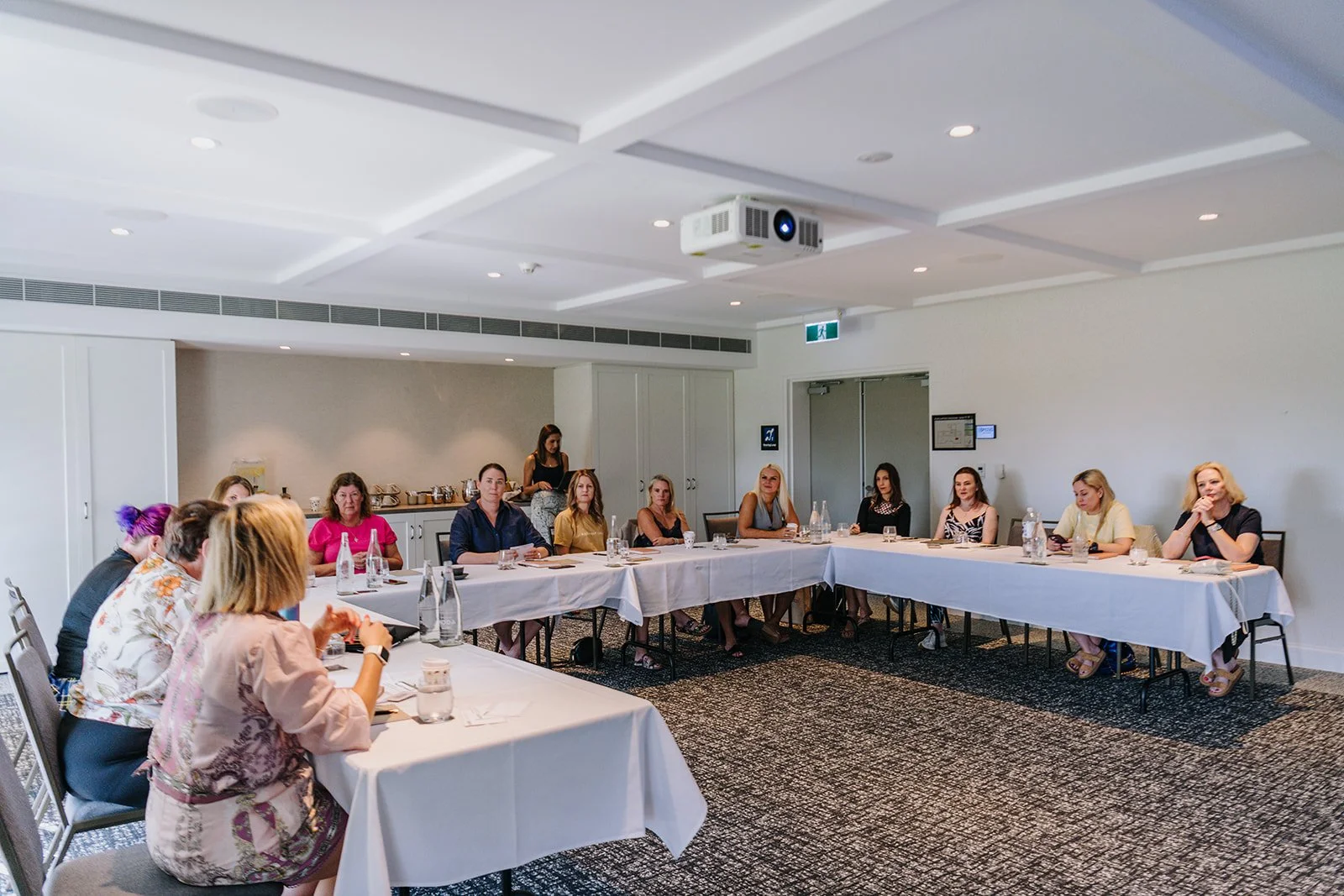 Several women seated around U-shaped conference table in a bright room with white walls, carpeted floor, and ceiling-mounted projector.