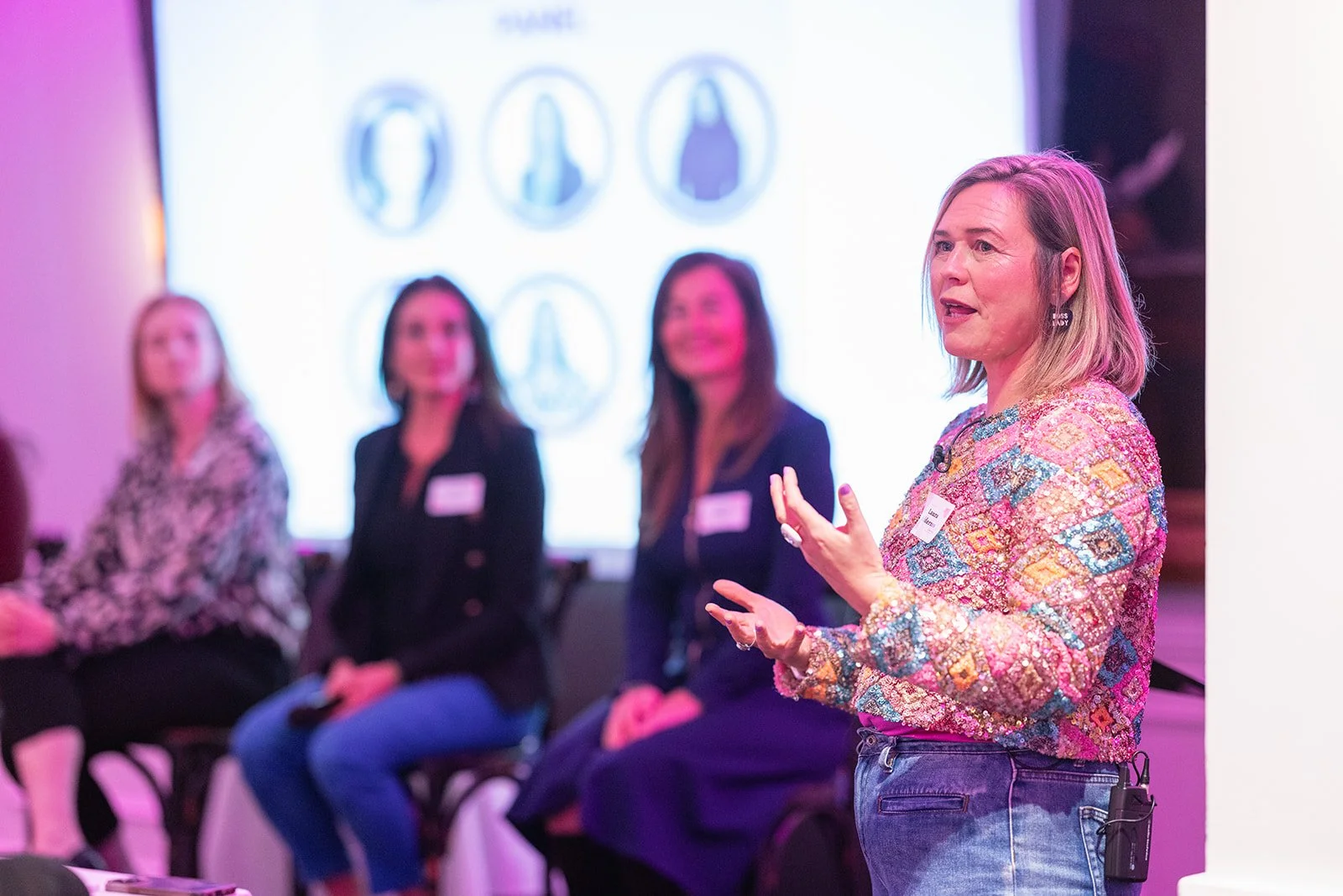 A woman in a colorful patterned blouse speaking to an audience during a panel discussion, with three women sitting behind her on stage and a large screen displaying silhouettes of four different earrings in the background.