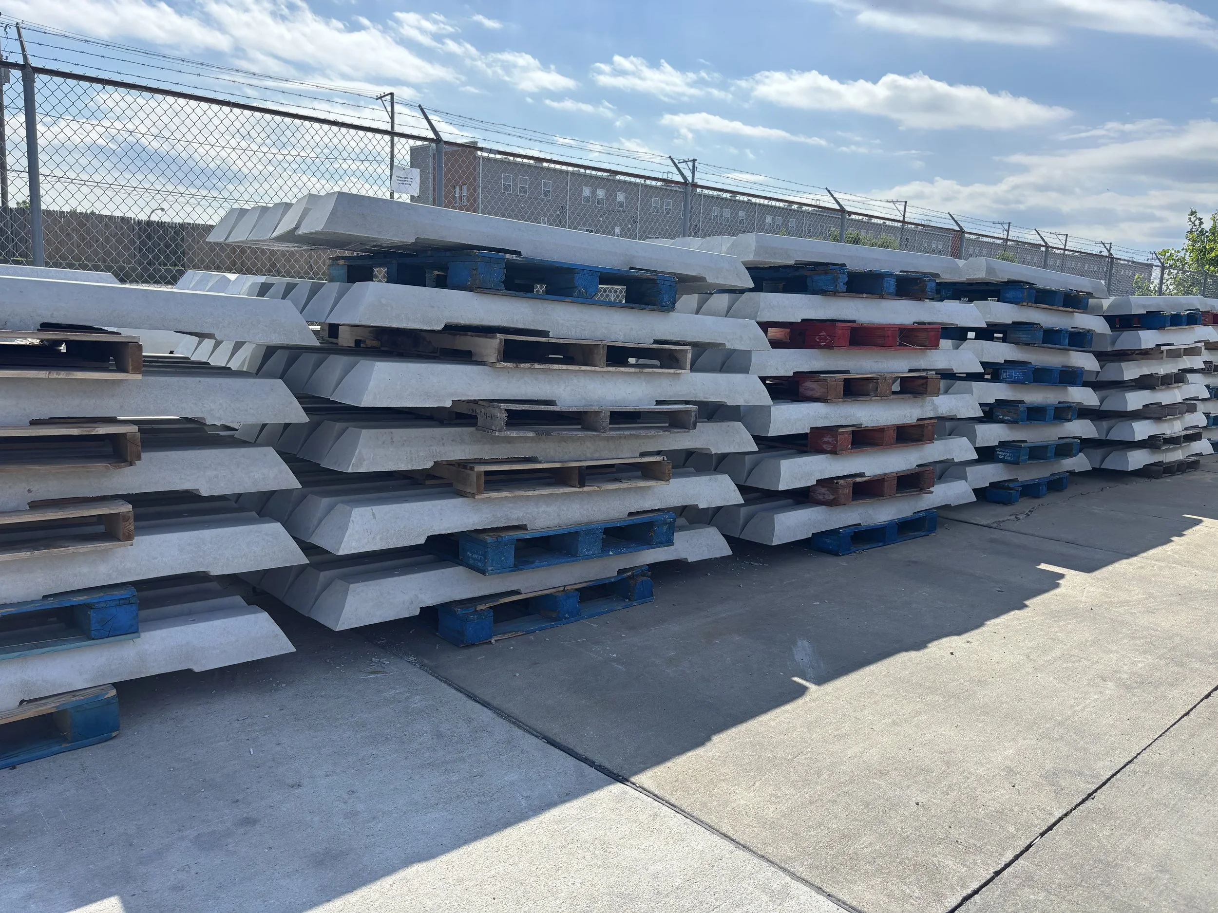 Stacked concrete curbs on blue wooden pallets outside, with a chain-link fence and buildings in the background under a partly cloudy sky.