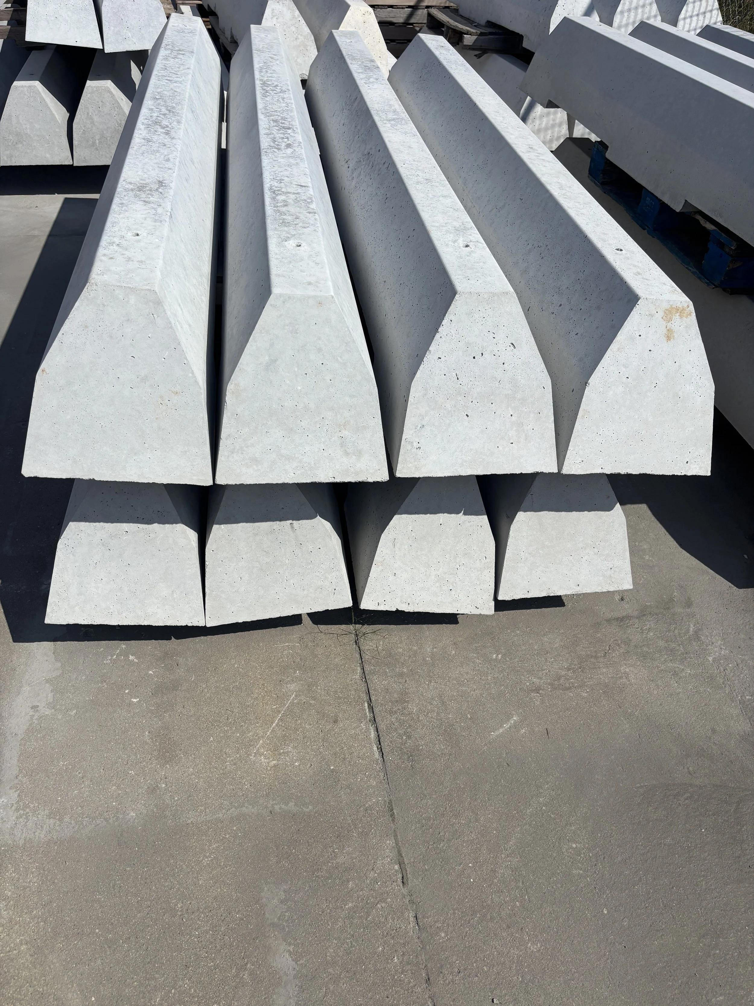 Stacked large concrete beams on a concrete surface with shadows cast on the ground.