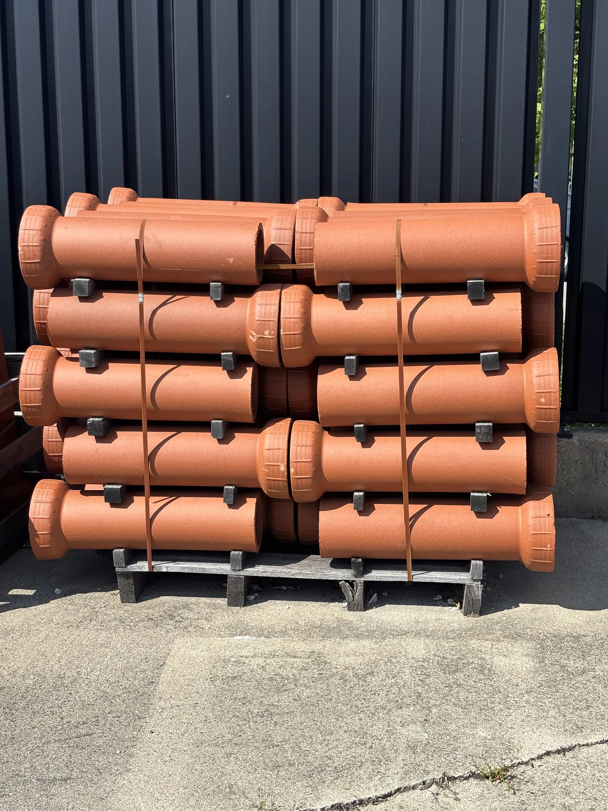 Stack of orange drainage pipes secured with straps on a wooden pallet outdoors near a metal fence.