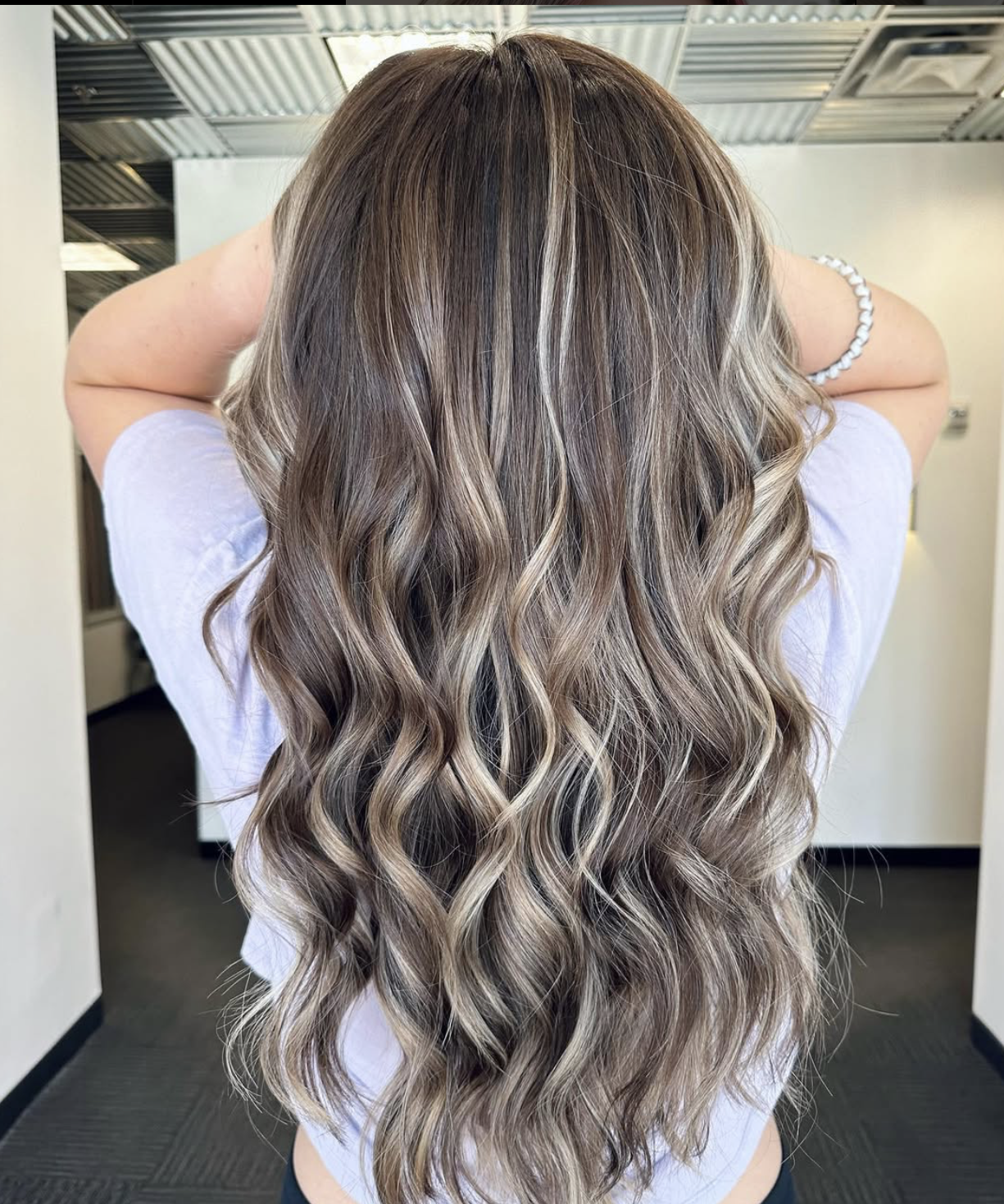 Woman with long, wavy, highlighted hair standing with her back to the camera, in an indoor setting.