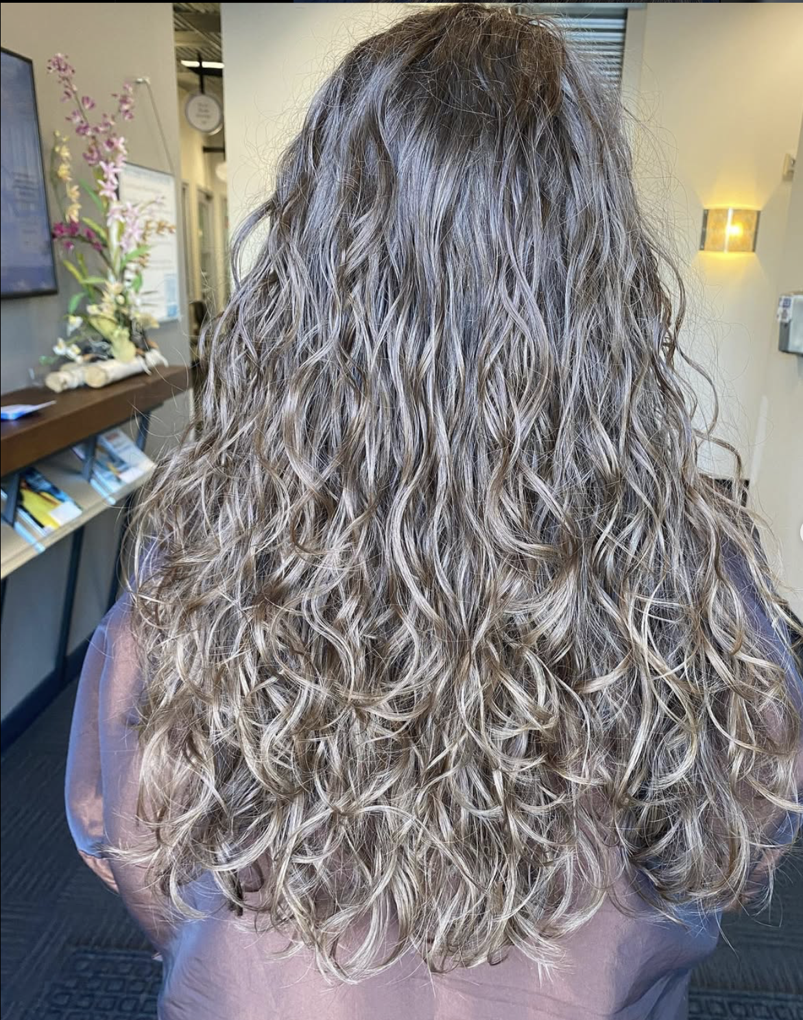 Back view of a woman with long, blonde, curly hair in a salon or waiting area.