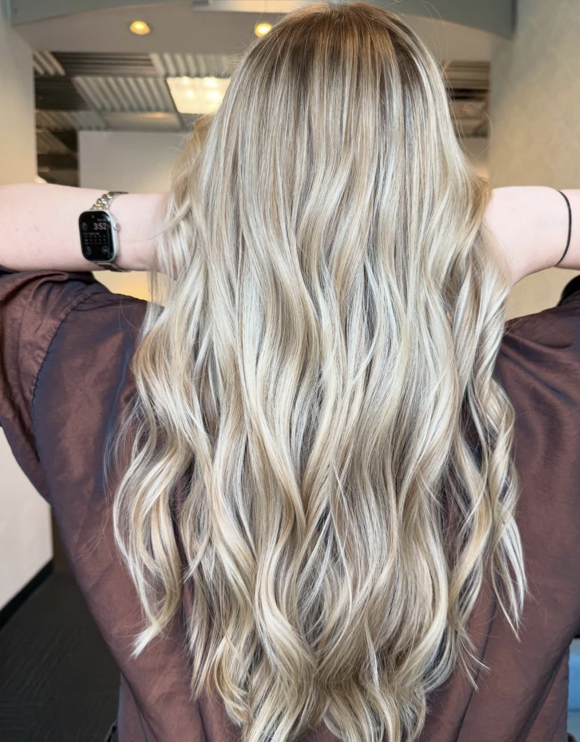 Back view of a woman with long, wavy blonde hair wearing a brown top and a smartwatch on her left wrist in an indoor setting.
