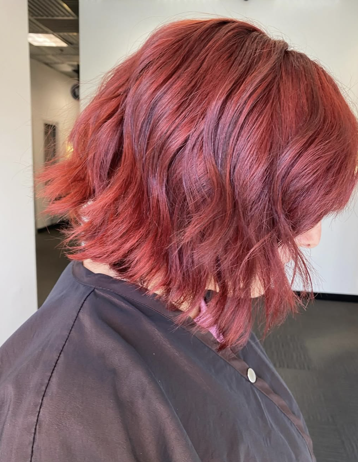 A person with shoulder-length, wavy red hair, seen from the side, inside a hair salon.
