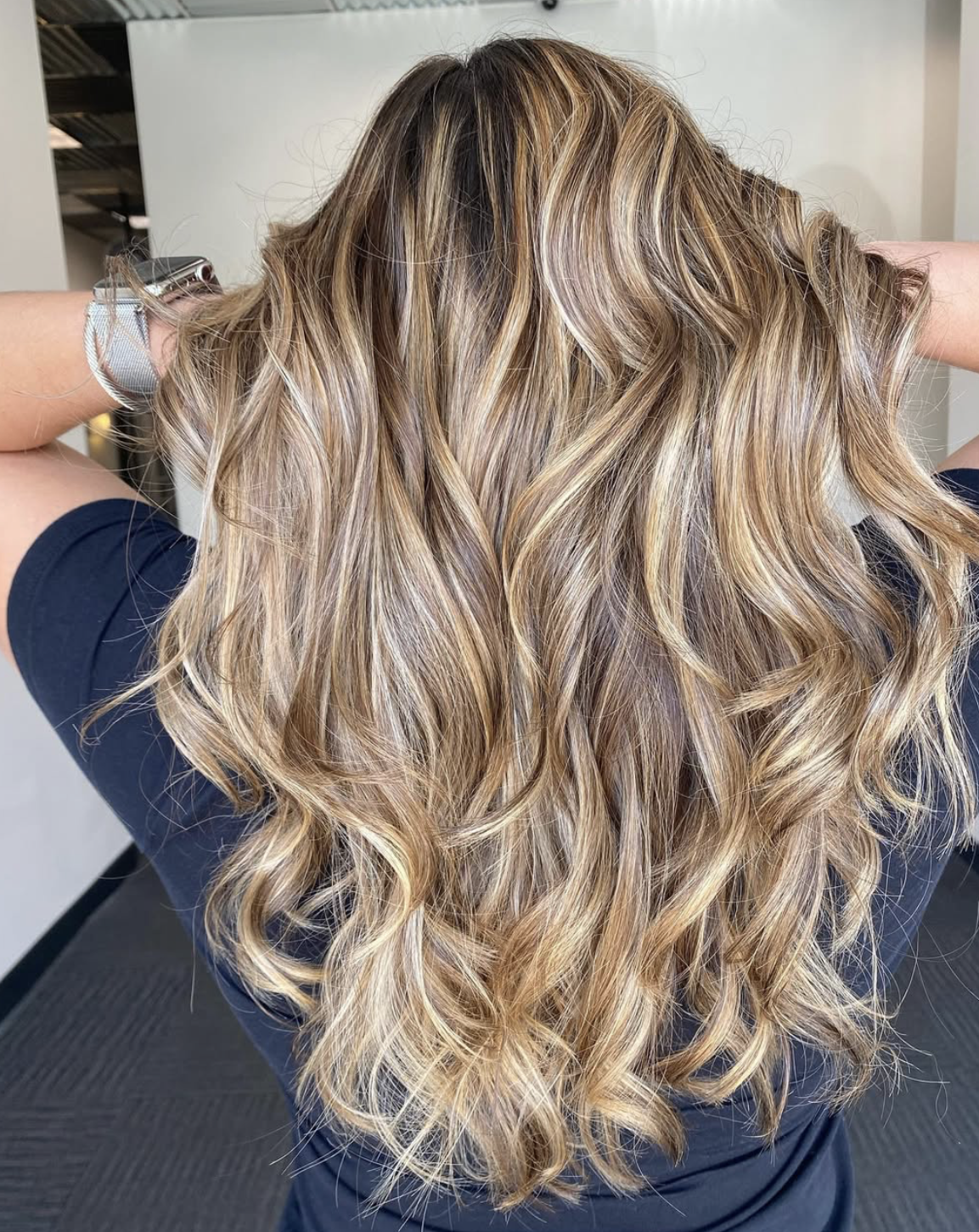 Woman with long, wavy blonde hair, wearing a dark shirt, viewed from the back.
