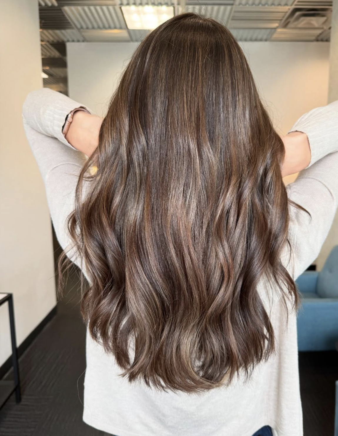 Back view of a woman with long, wavy, brunette hair with subtle highlights, wearing a light-colored sweater, standing indoors.