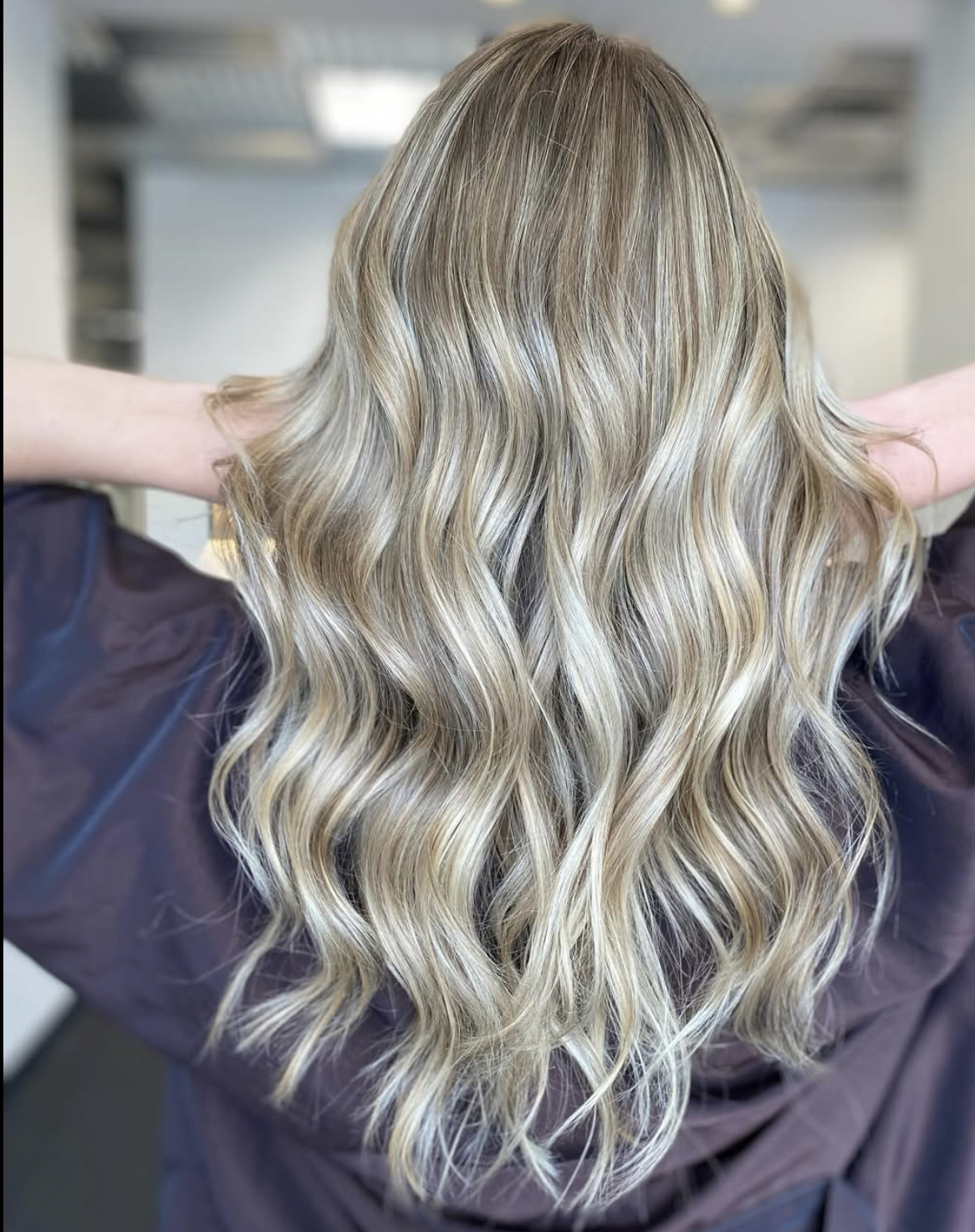 Back view of a woman with long, wavy blonde hair, seated in a salon chair with arms raised, wearing a dark cape.