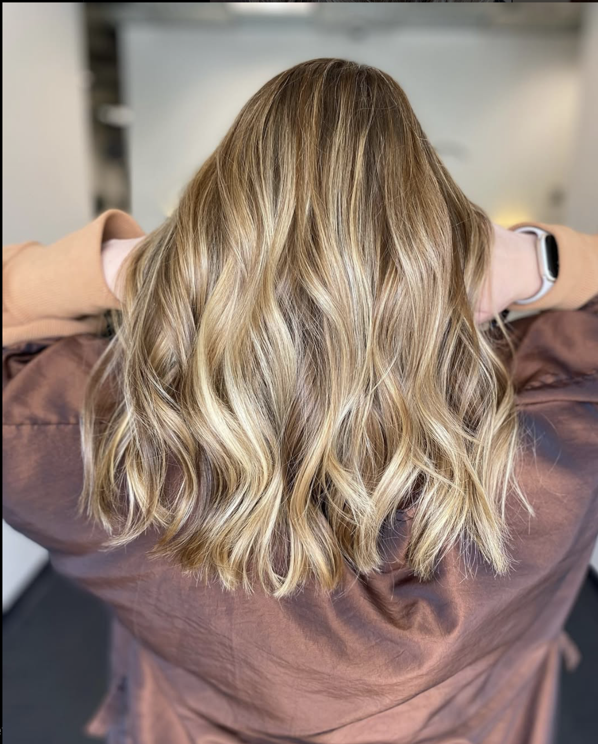 Back view of a woman with long, wavy blonde hair wearing a brown salon cape and a smartwatch.