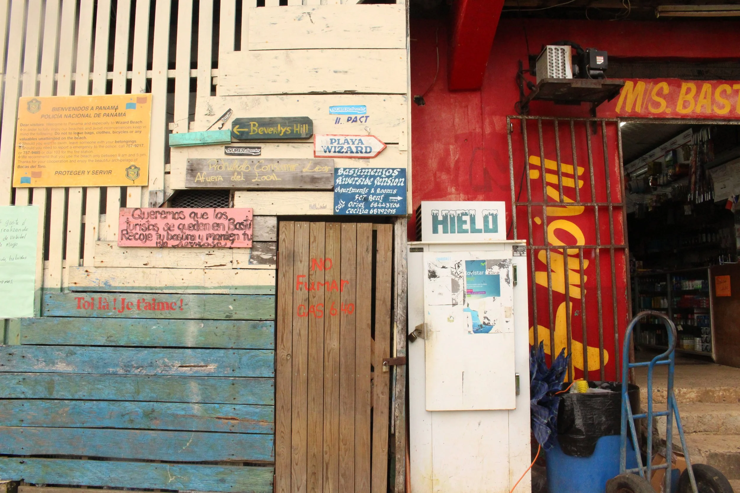 A wall with various signs, including one in Spanish for tourists about staying in Basti, a sign for a local waterfall, signs for Playa Wizard and Basamentos Riverside, a blue sign for ice, and a wooden door that says 'No fumar, GAS 640'. There is als