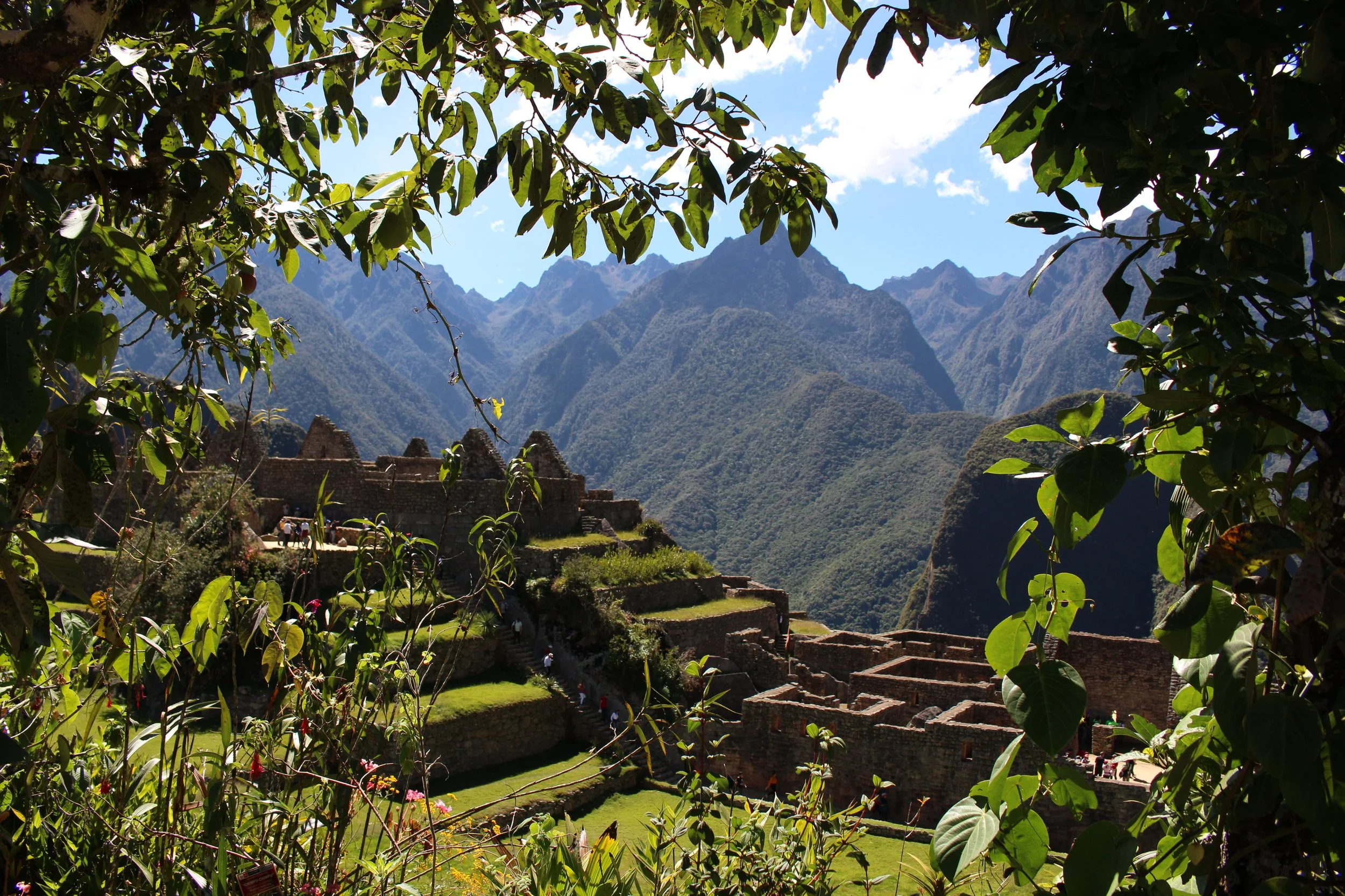 Ancient Incan ruins surrounded by lush green vegetation, with mountainous terrain in the background under a partly cloudy sky.