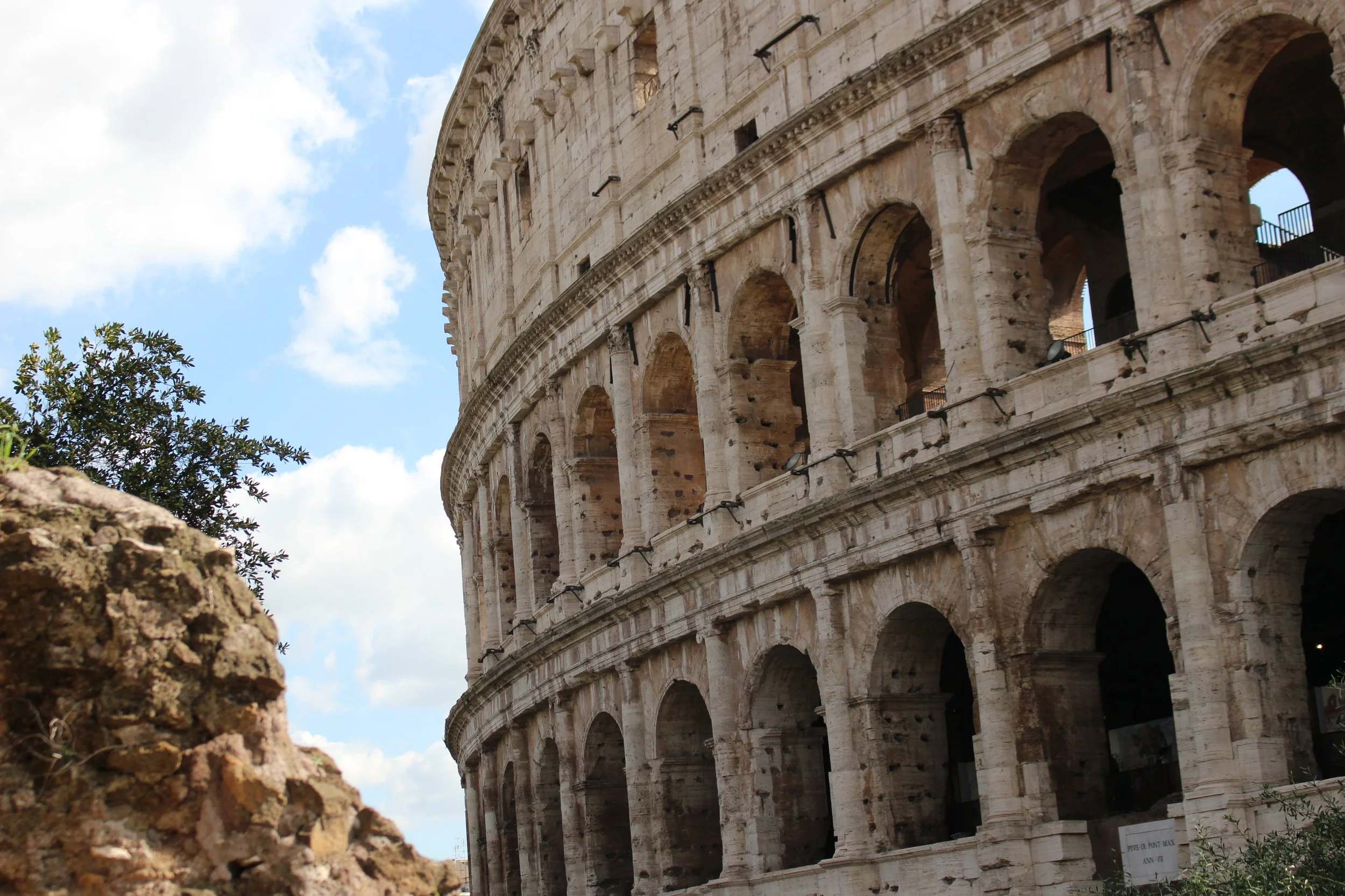 A close-up view of the Roman Colosseum showing its ancient stone arches and columns with a cloudy blue sky in the background.