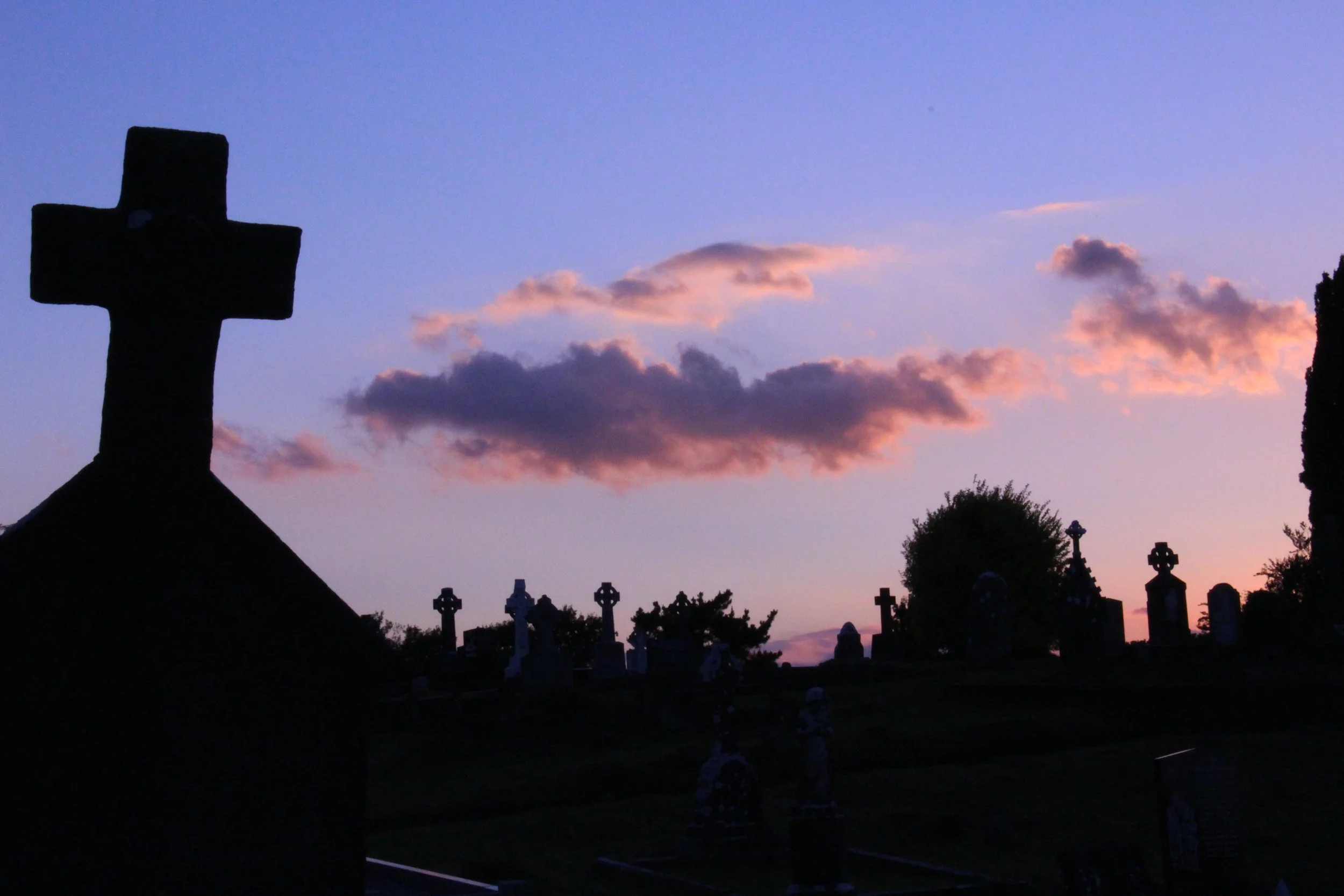 Silhouette of a large cross monument in a graveyard at sunset with clouds in the sky.