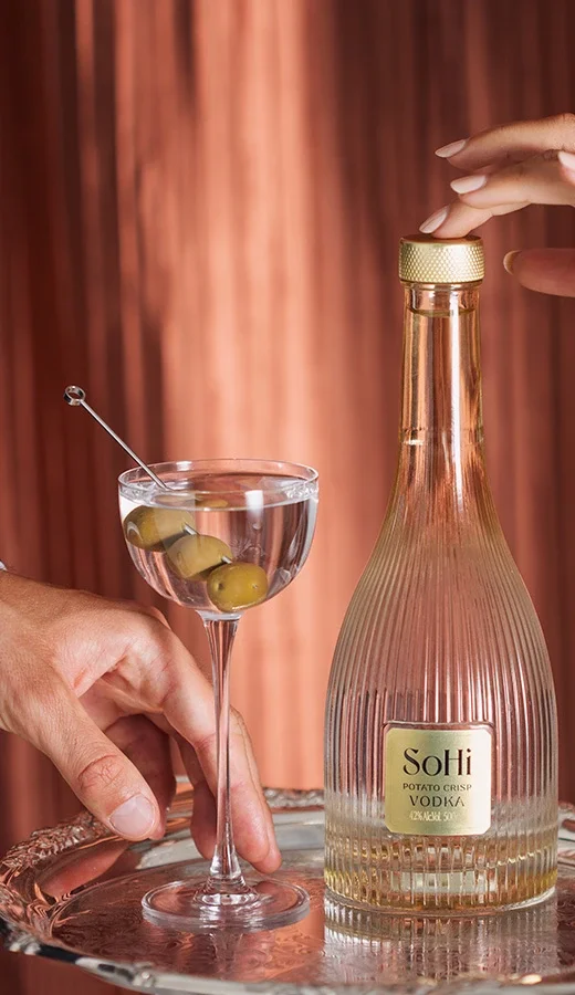 A hand holding a martini glass with green olives and a cocktail pick, and another hand opening a bottle of SoHi vodka on a reflective tray with a copper-colored background.