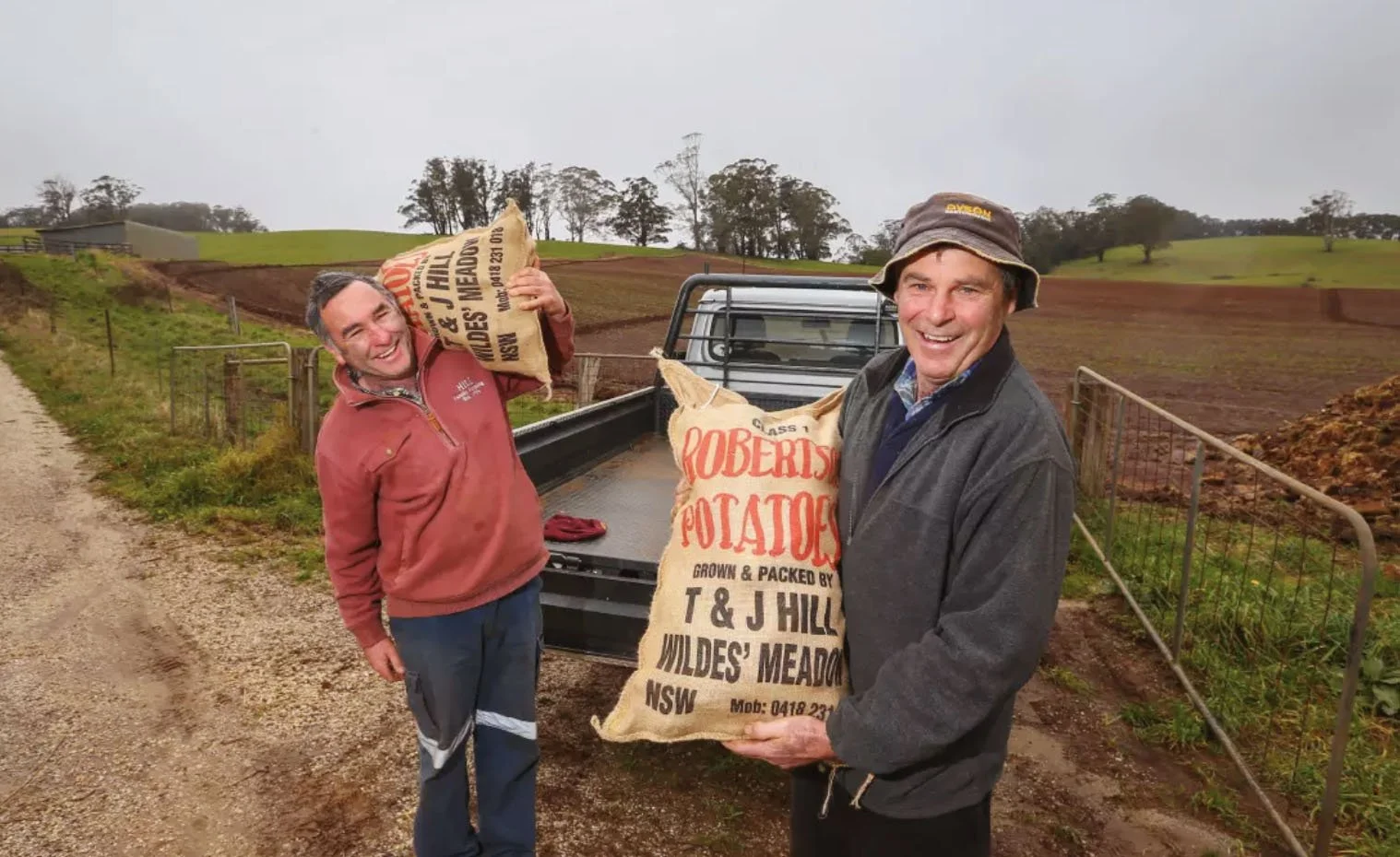 Two men on a farm holding bags of potatoes, standing next to a pickup truck with a rural landscape in the background.