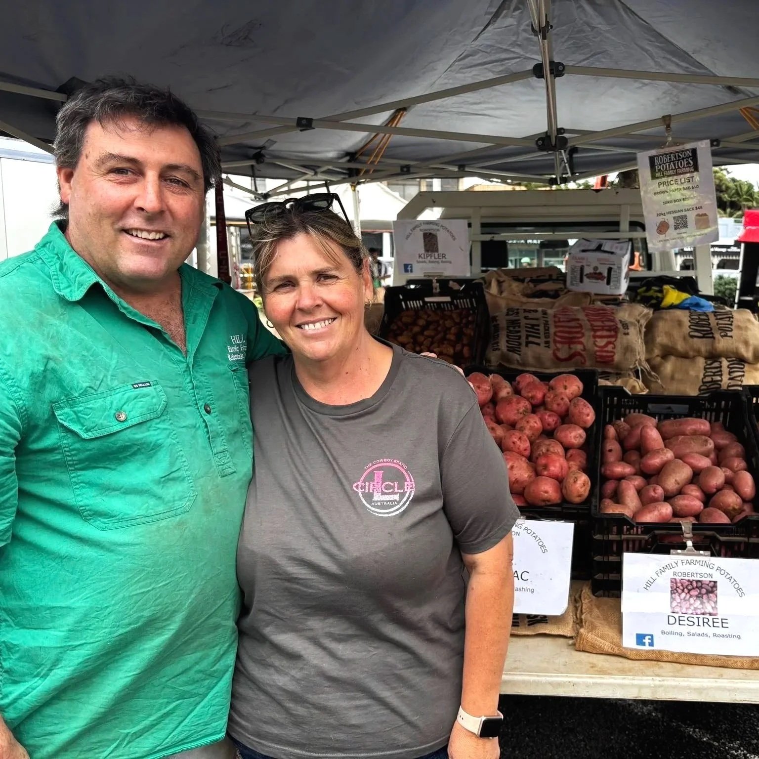 A man and woman smiling at a farmer's market stall with potatoes and signage in the background.