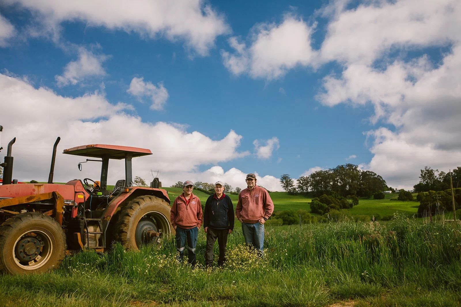 Three men standing in a grassy field next to a red tractor on a farm, with green rolling hills and trees in the background under a partly cloudy sky.