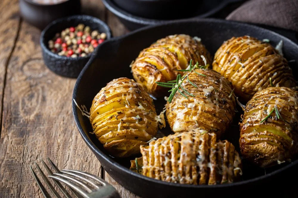 Baked Hasselback potatoes garnished with rosemary in a black skillet, with a small bowl of mixed peppercorns in the background.