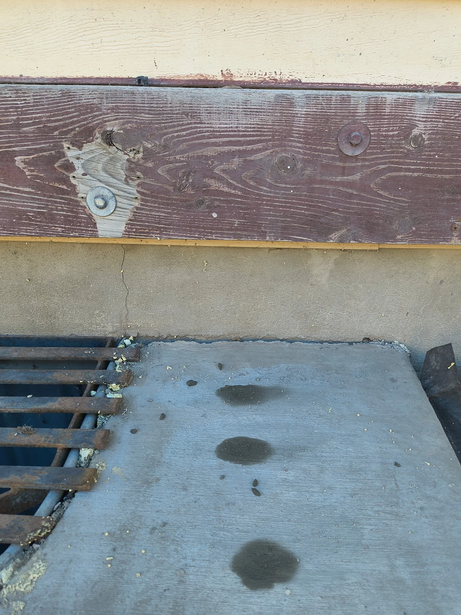 Close-up of a concrete step with water spots and a metal grate on the left. The background shows a wooden board attached to a wall.