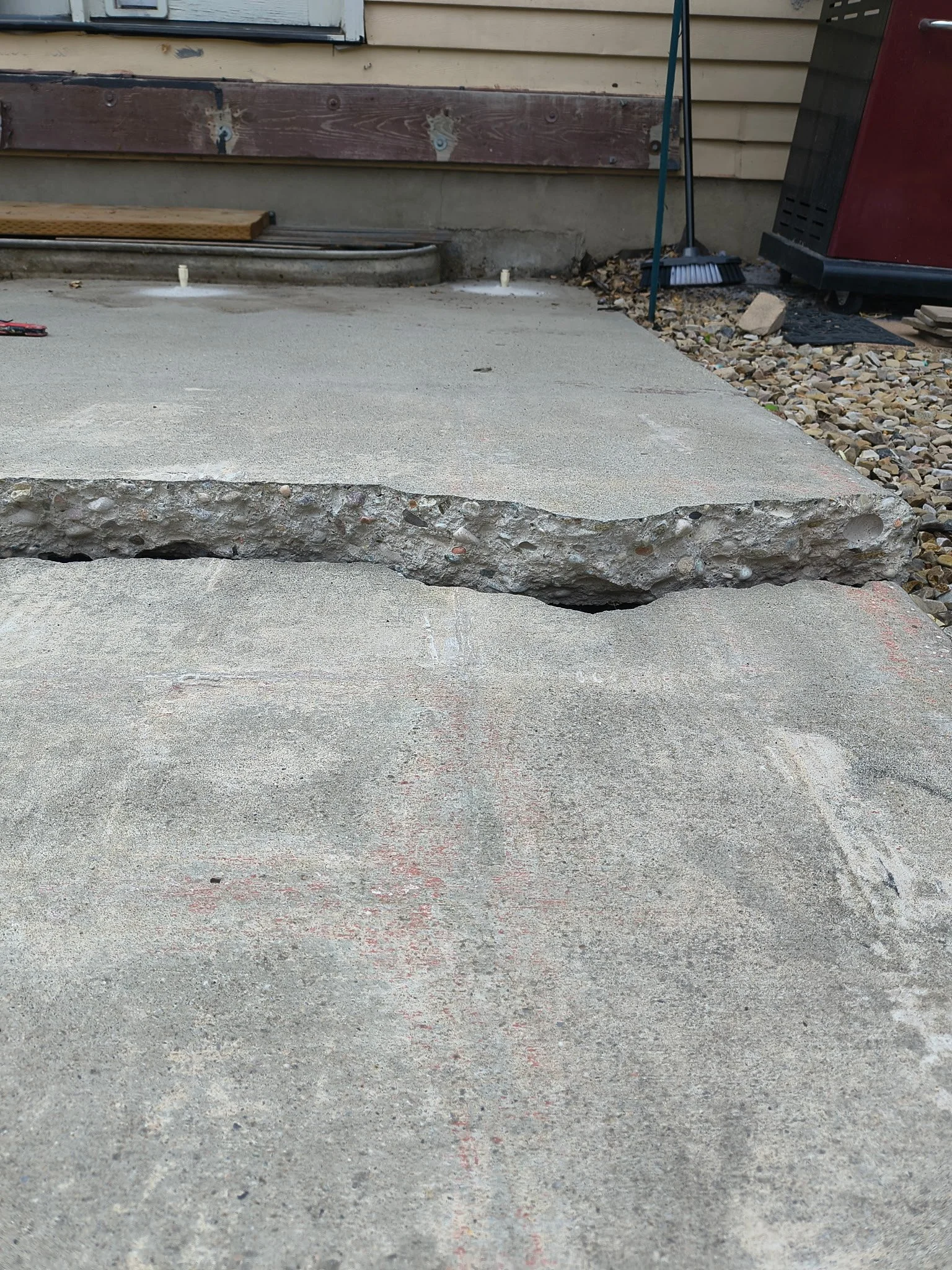 Cracked concrete porch with a significant horizontal break in the middle, next to a house with beige siding, a rake, a grill, and gravel on the side.