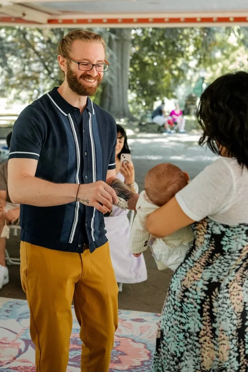 Man using sage to bless baby during baby naming ceremony who is being held by mother in outdoor setting in Prospect Park