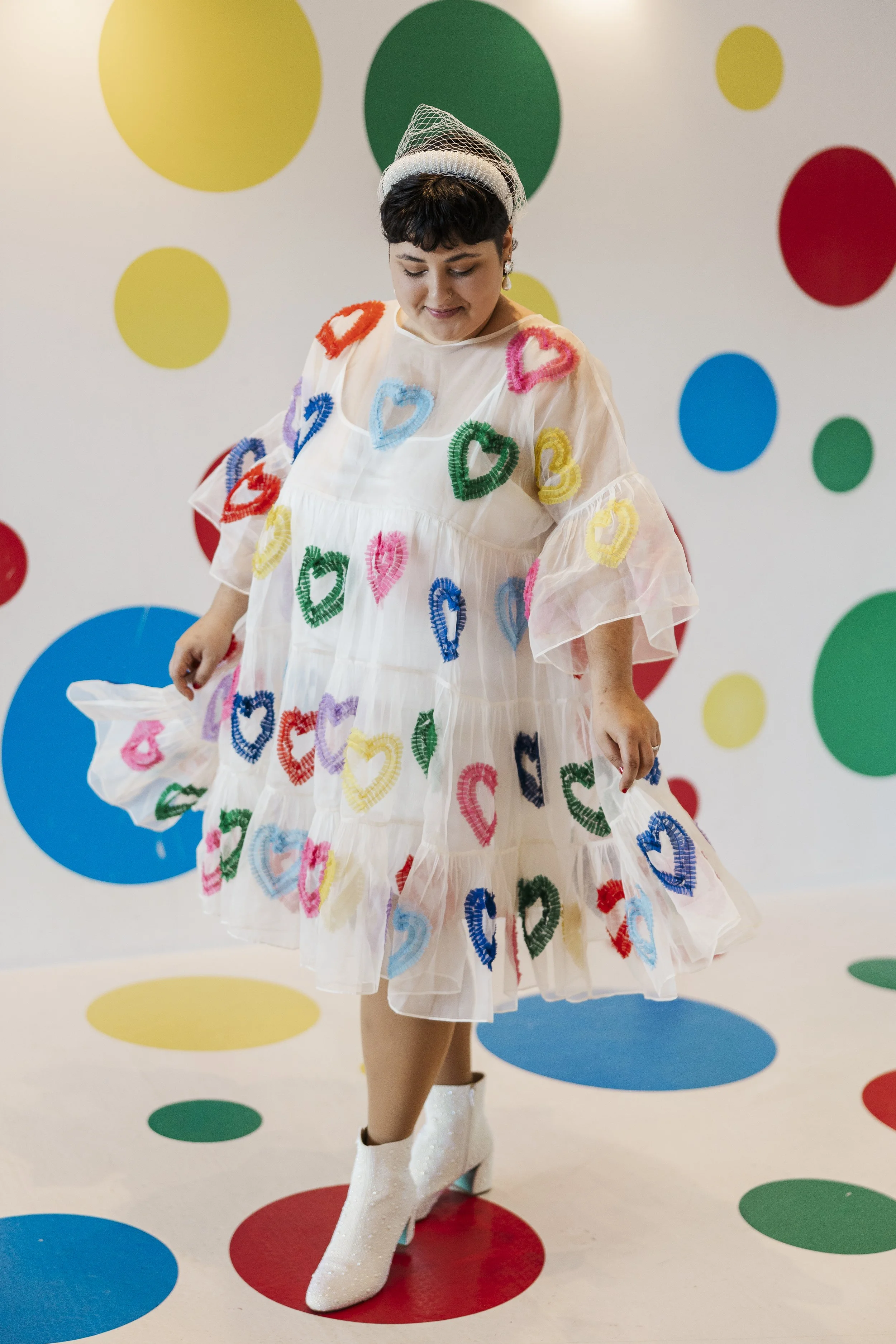 Woman in multi-color heart dress looking down at her dress with a polka dot wall behind her, image for a self-love ceremony