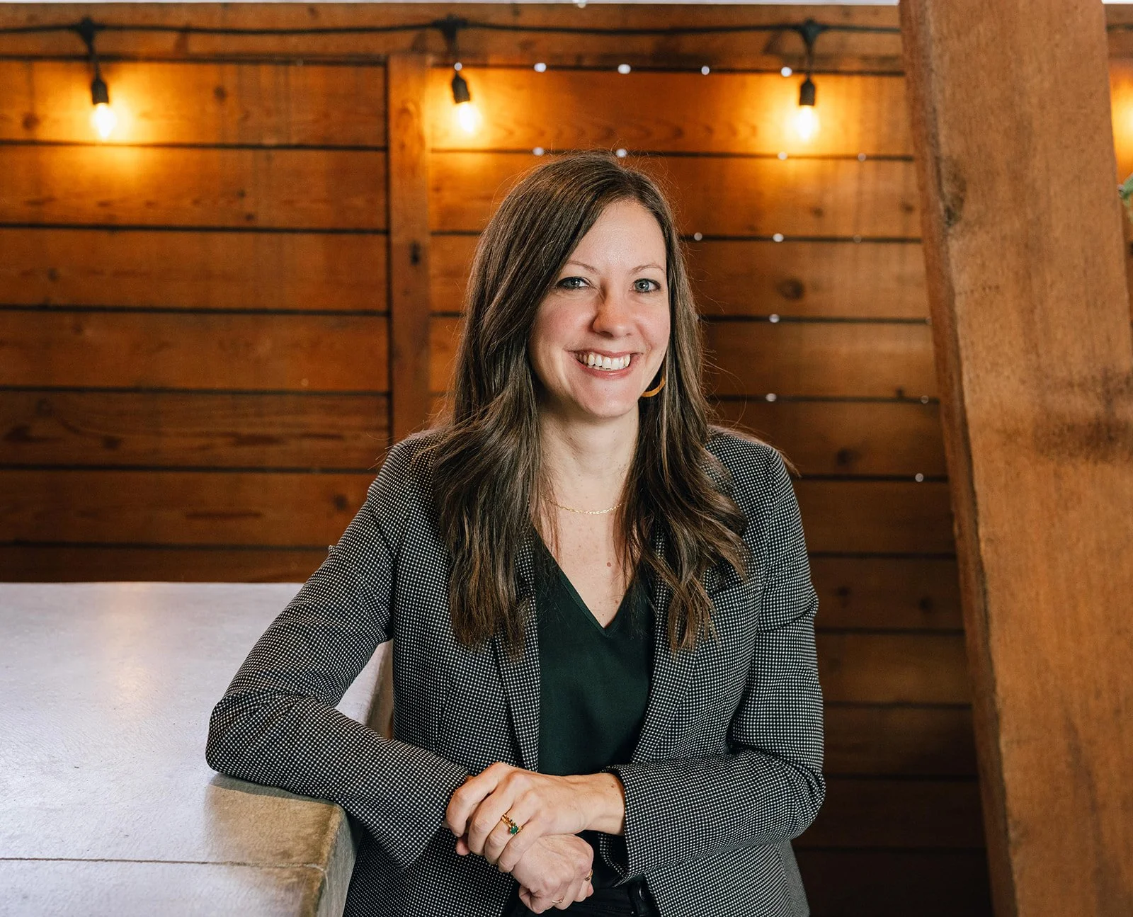 A woman with long brown hair and light skin sitting at a table, smiling. She is wearing a black top and a gray blazer with a checkered pattern in an indoor setting with wooden wall panels and warm lighting.