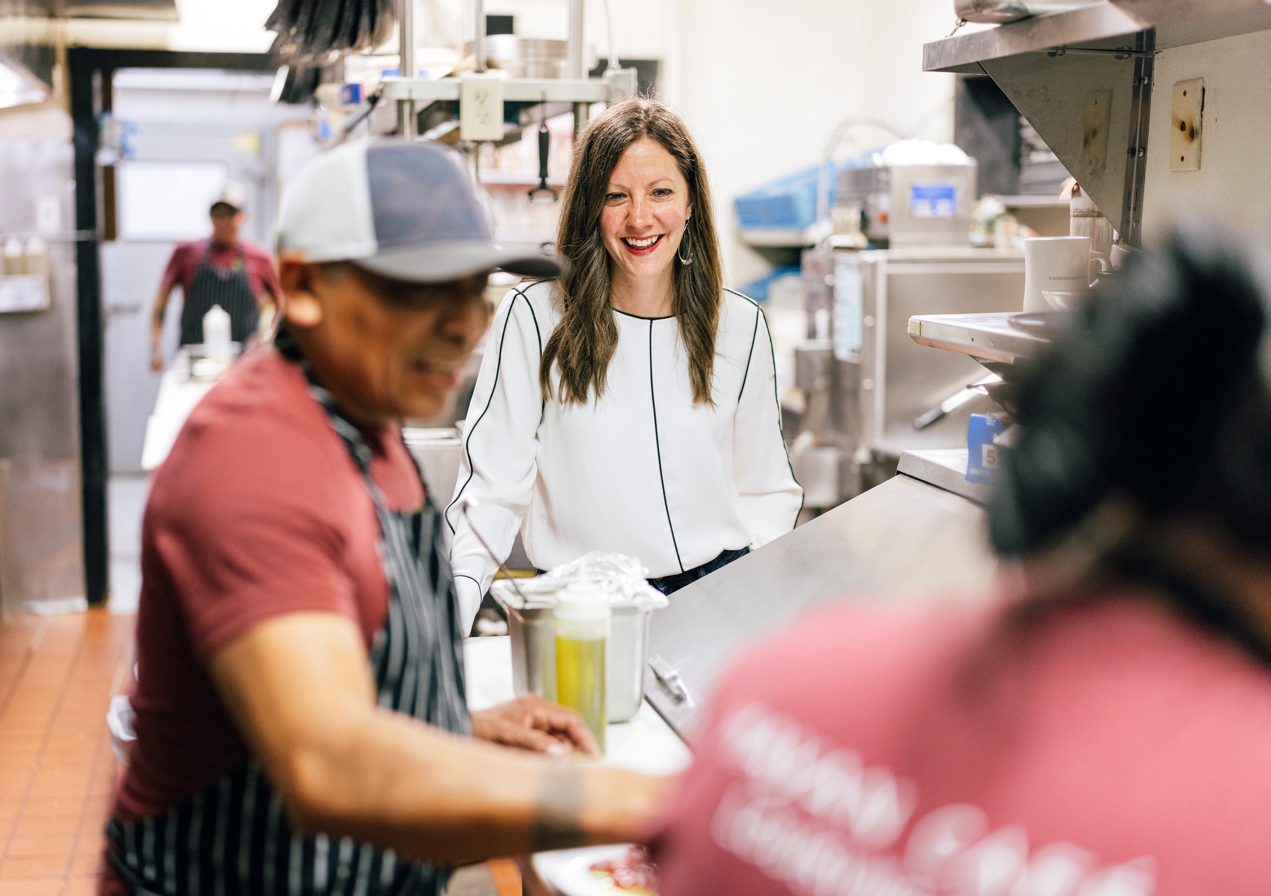 A woman with long brown hair, smiling, standing in a busy kitchen or cafeteria, wearing a white top with black piping. The image also shows a person in a red shirt and striped apron in the foreground, and another person in the background wearing a red shirt and black apron. The setting appears to be a commercial kitchen with shelves, appliances, and food-related equipment.