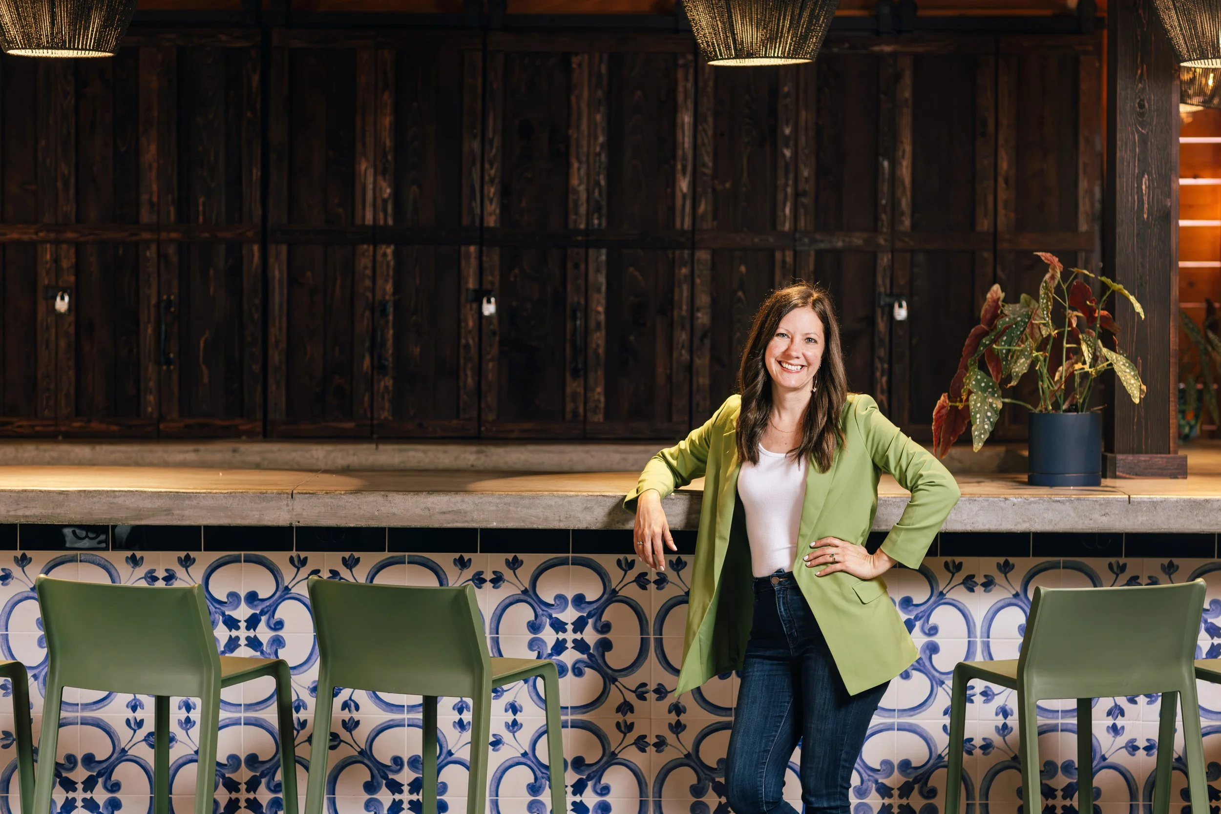 A woman with long brown hair smiling, wearing a lime green blazer and white shirt, standing in front of a bar counter with green chairs and patterned tiles, in a warm-lit restaurant or bar with dark wood and decorative lighting.