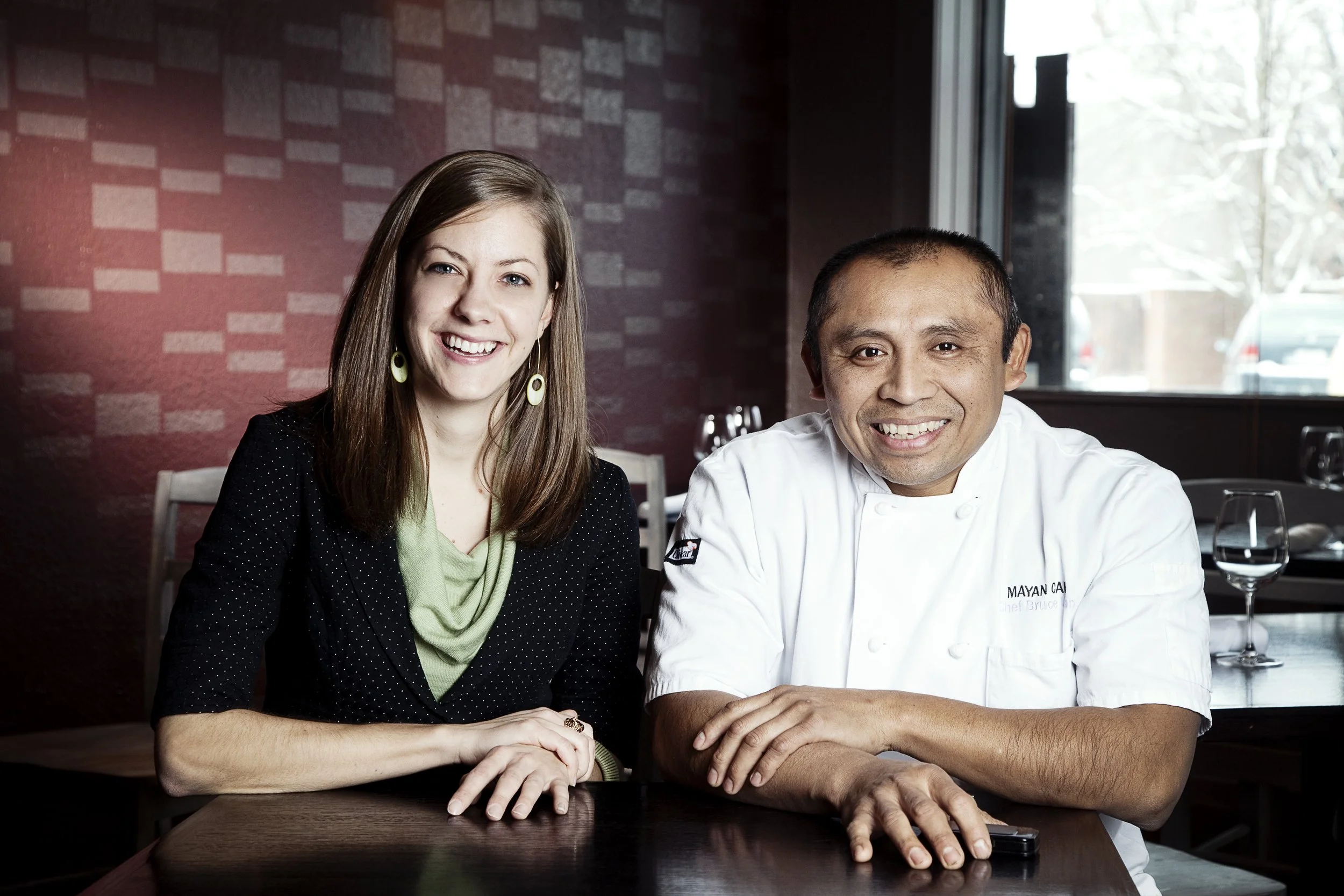 A woman with long brown hair, wearing a black jacket and green top, sitting at a table next to a smiling man in a white chef's coat. They are in a restaurant with a red brick wall and a window in the background.