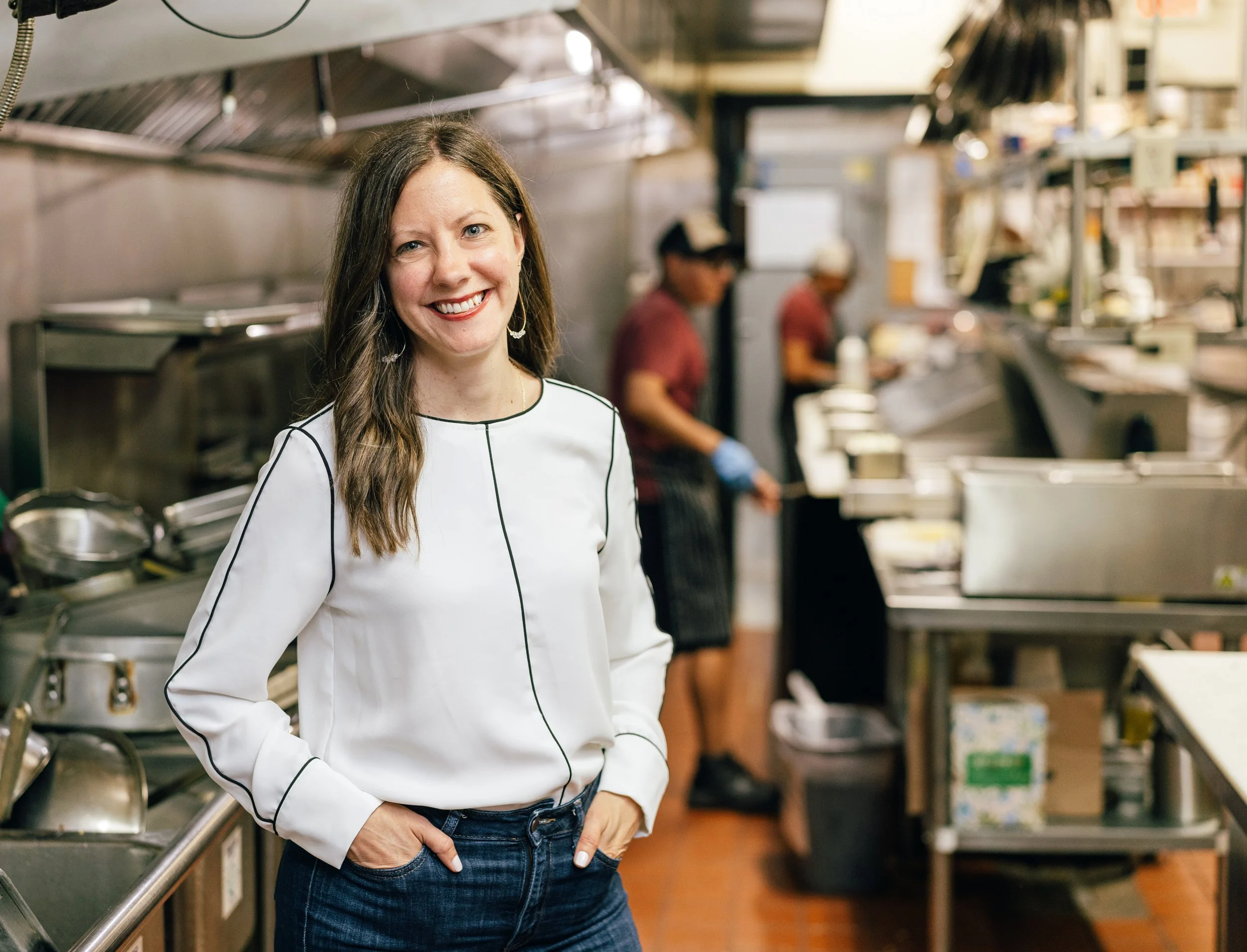 Woman smiling in a professional kitchen with two other people focusing on food preparation in the background.